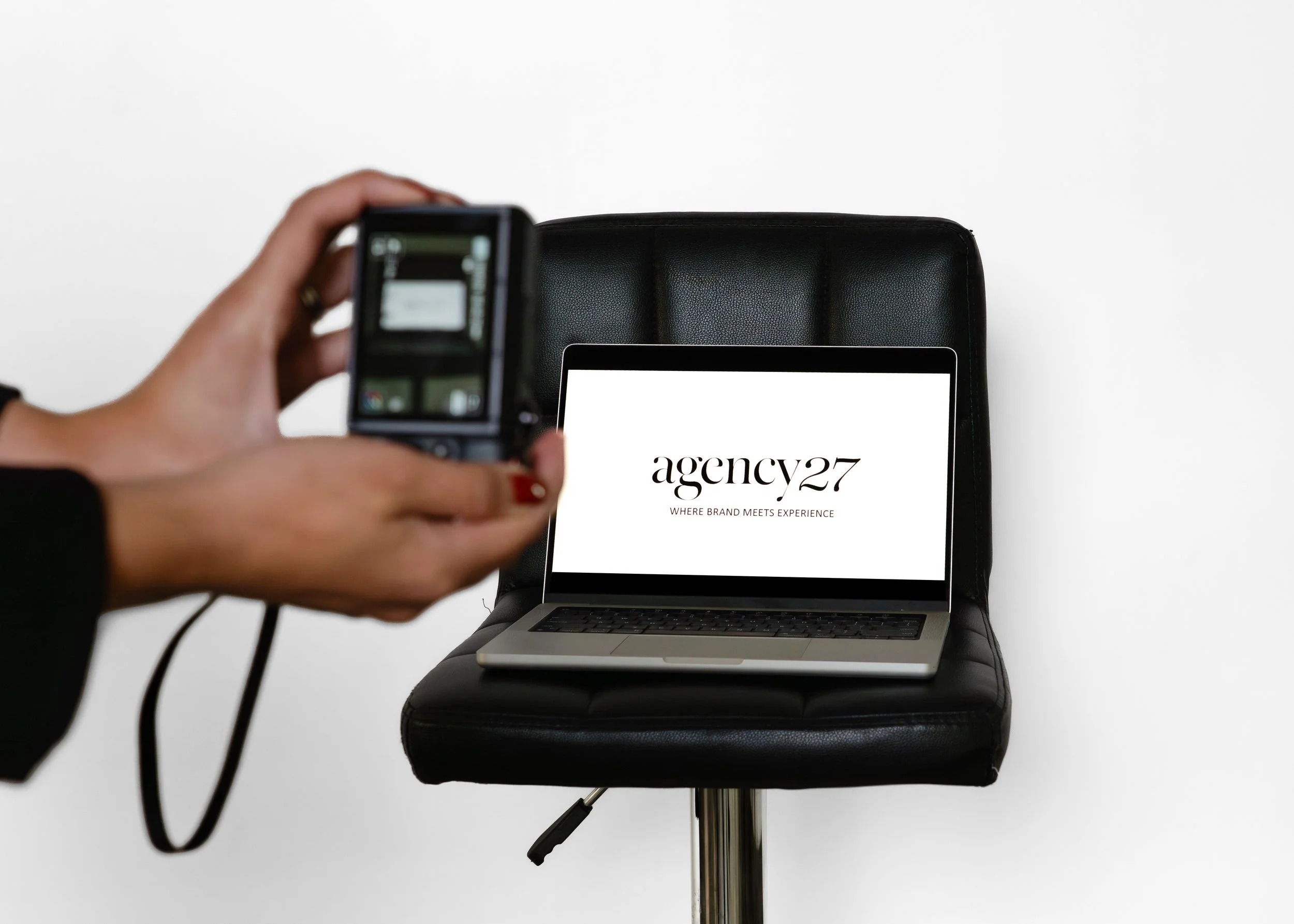 Person taking a photo of a laptop displaying the logo 'agency27' with the tagline 'WHERE BRAND MEETS EXPERIENCE,' on a black chair against a white background.