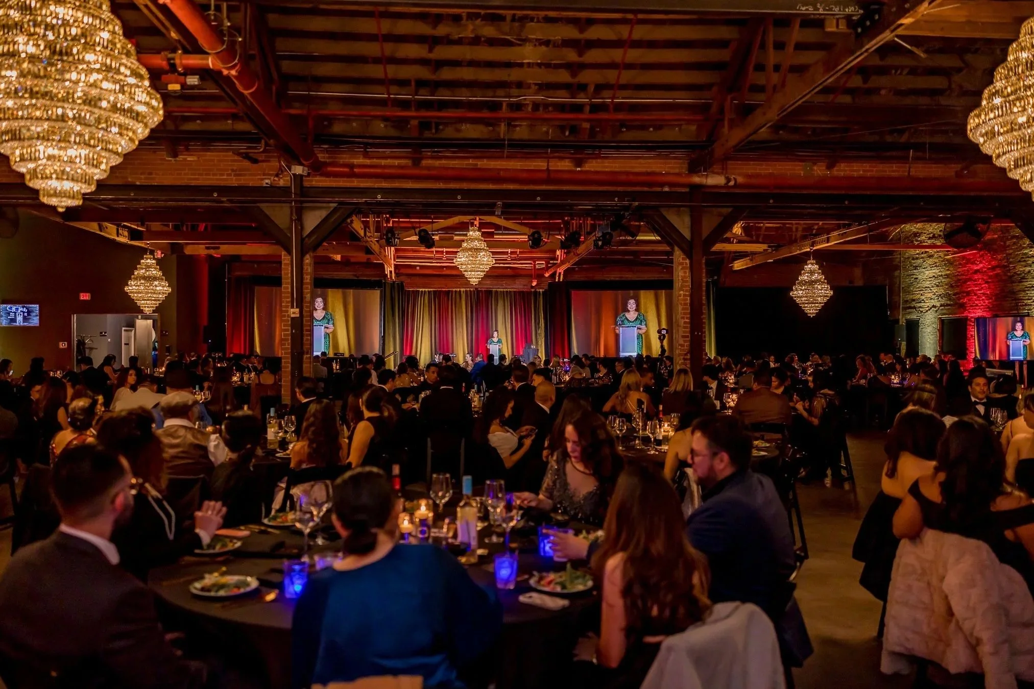 People attending a formal event or dinner in a dimly lit venue with chandeliers, a stage with a speaker, and decorated tables.