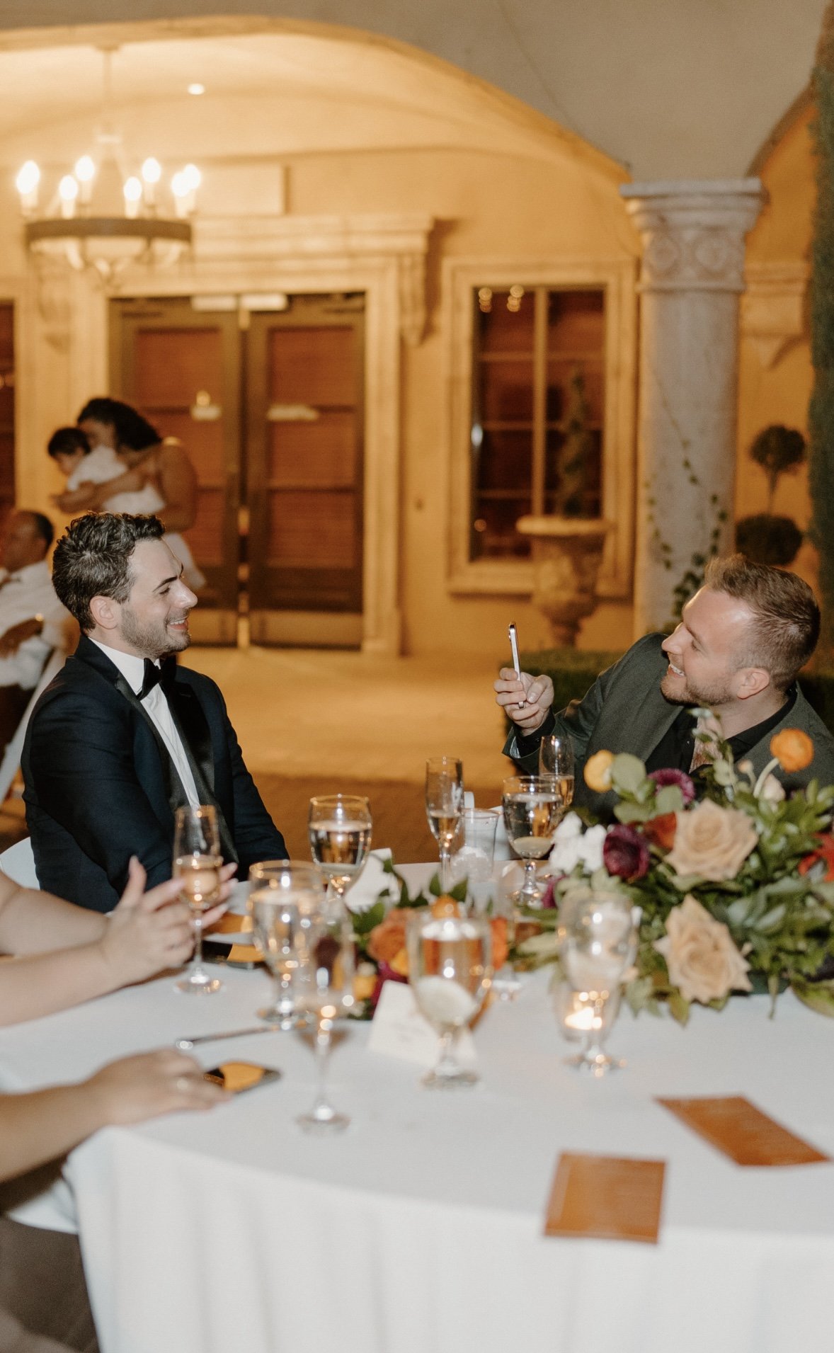 Two men sitting at a formal dinner table, smiling and taking a selfie, with a floral centerpiece and glasses of wine, in a warmly lit indoor setting.