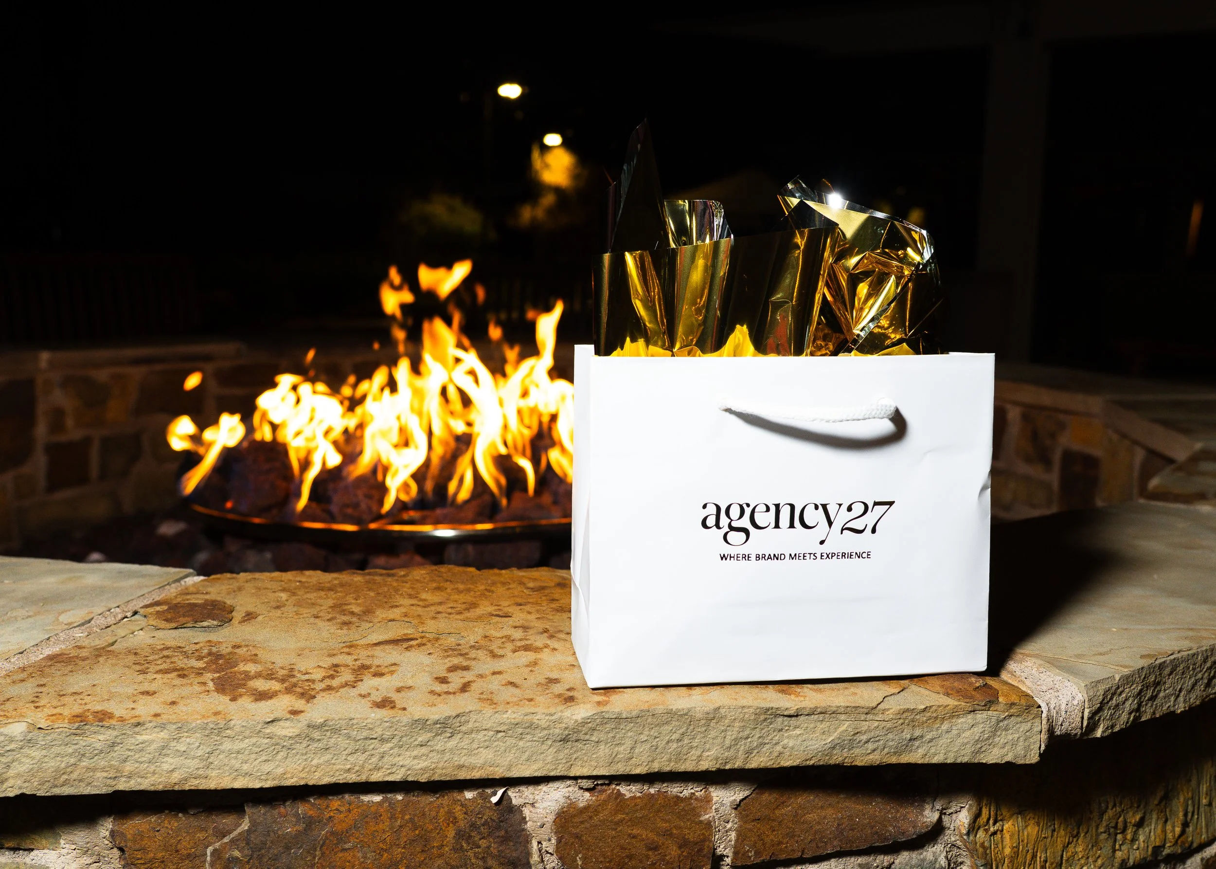 White shopping bag with black text and rope handle on stone surface in front of a fire pit with visible flames at night.