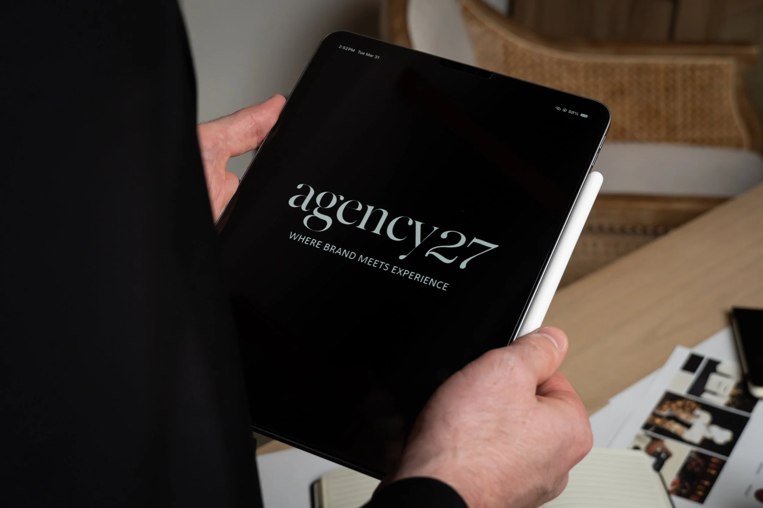 A person holding a tablet displaying a black screen with the text 'agency27' and the tagline 'Where Brand Meets Experience'.