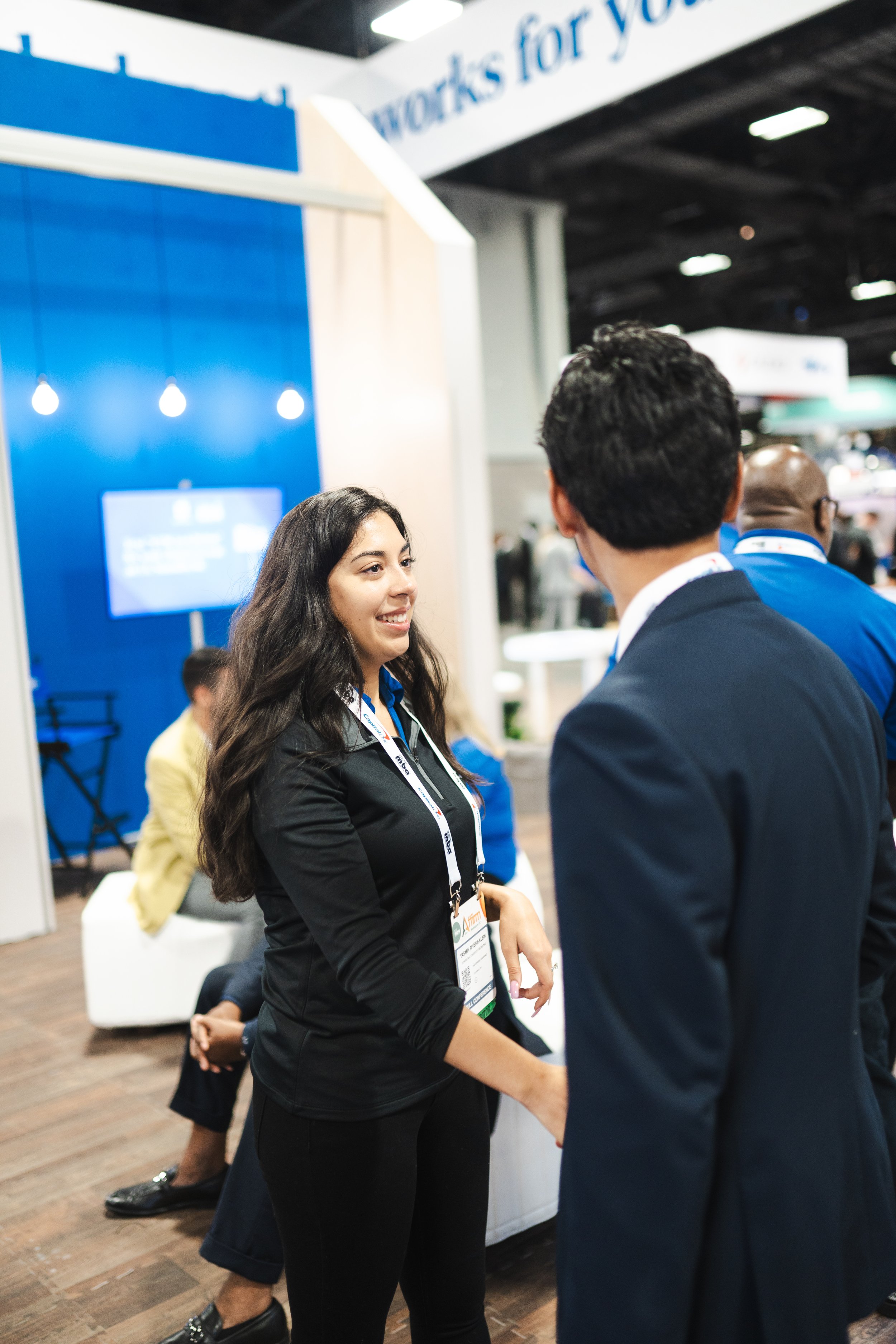 A woman and a man shaking hands at a trade show or conference, with other attendees in the background and a blue booth behind them.
