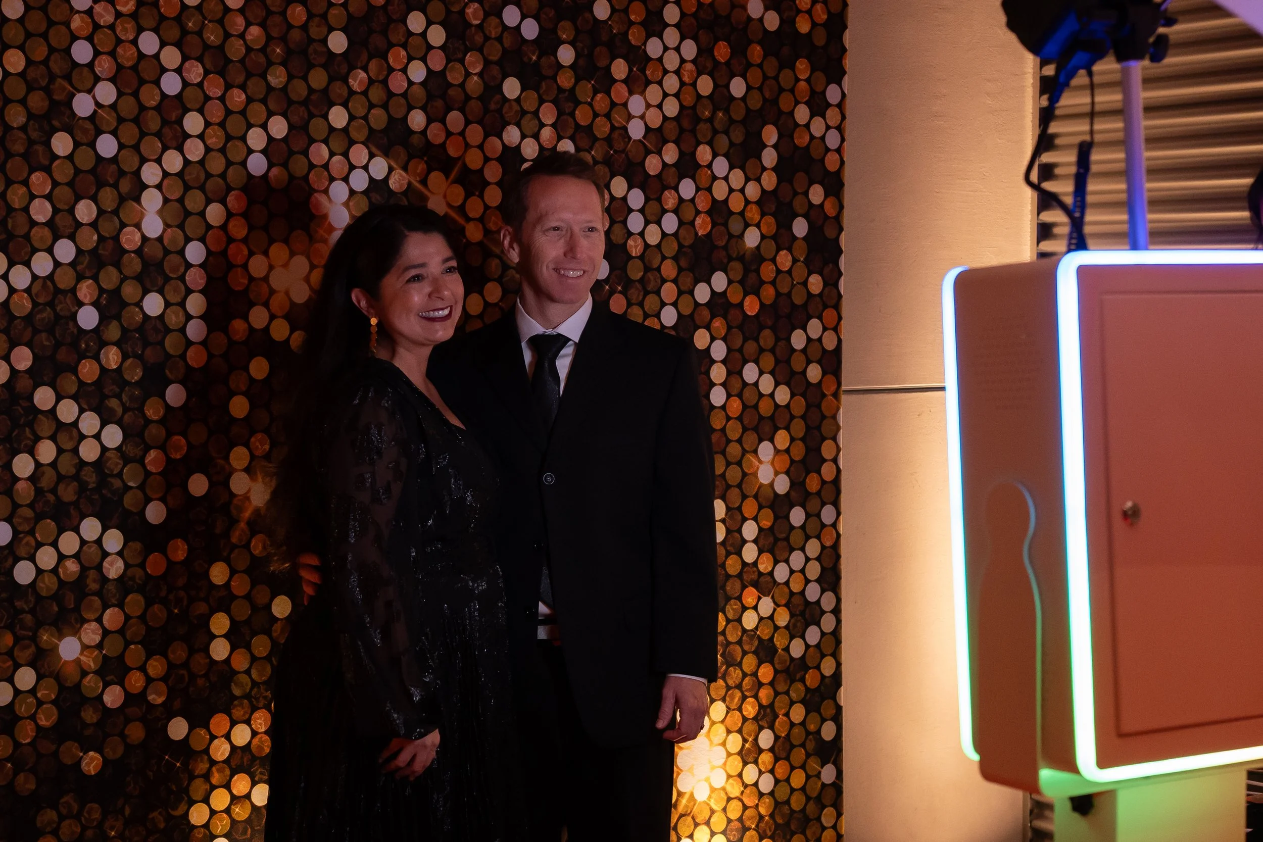 Two people, a woman and a man, dressed in formal attire, standing and smiling in front of a decorative background with circular lights. There is a bright, colorful illuminated photo booth on the right side of the image.