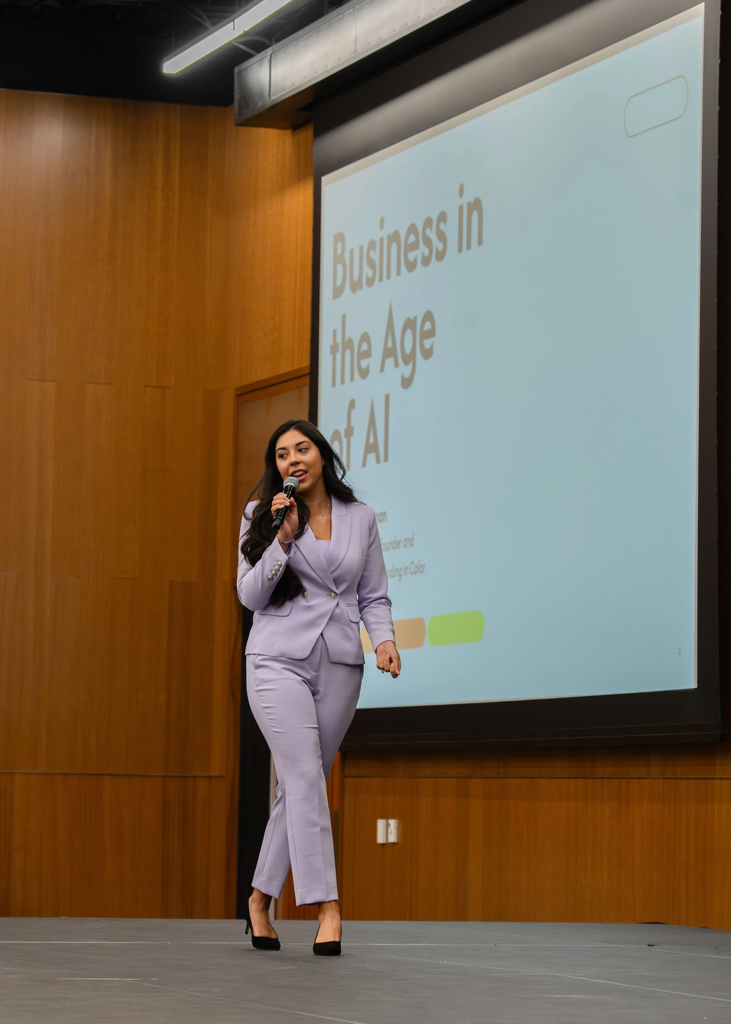 A woman in a lavender suit speaking on stage at a conference, with a presentation slide titled 'Business in the Age of AI' projected behind her.