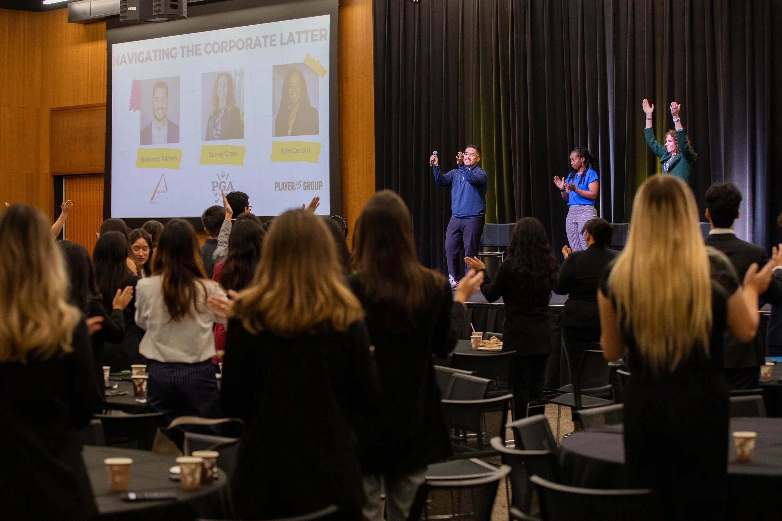 A large group of people attending a conference or seminar, with some on the stage and most standing and raising their hands. The stage has a screen displaying a presentation with the title 'Navigating the Corporate Latter' and pictures of three speakers. The setting appears professional and the audience is engaged.