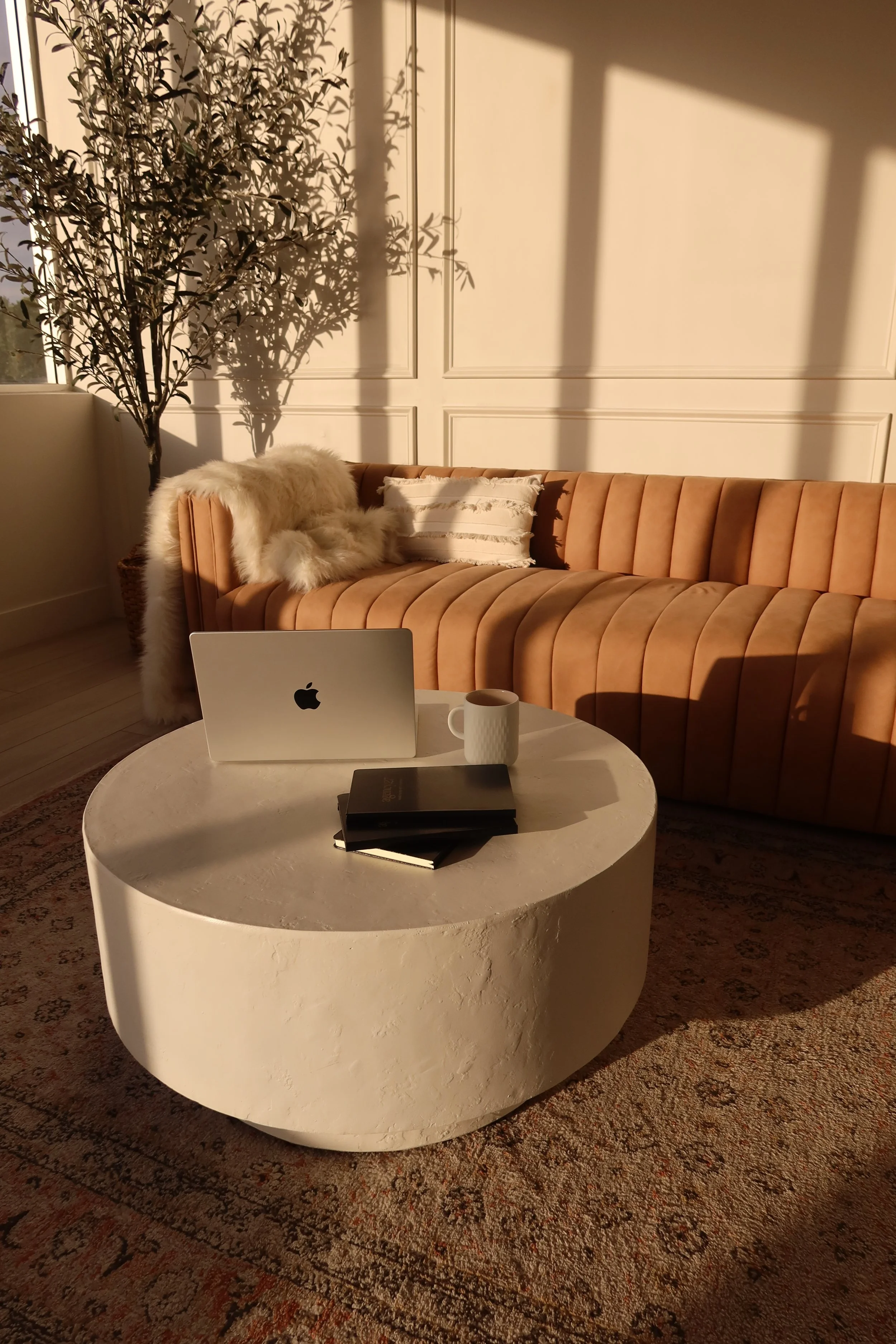A cozy living room corner with a salmon-colored velvet sofa, a white marble round coffee table with an Apple MacBook, a white mug, and a closed black notebook on it. Sunlight casts shadows on the wall, illuminating a tall plant in a basket and a white cushion with fringed edges on the sofa, all resting on a patterned rug.
