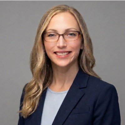 Professional headshot of a woman with long blonde hair, glasses, and a black blazer, smiling against a plain gray background.