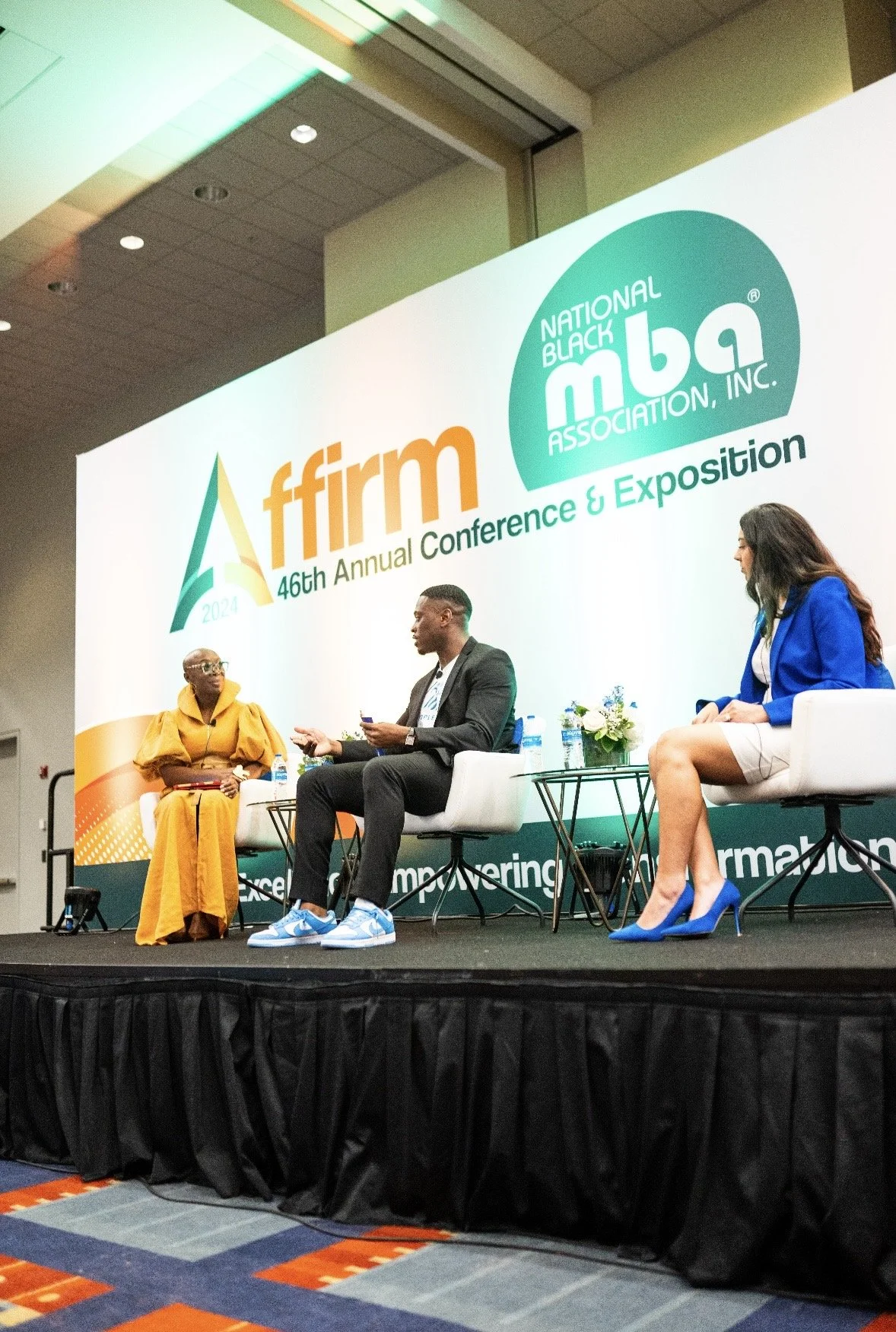 Three people sitting on a stage at the AFFIRM 46th Annual Conference and Exposition, with a large backdrop displaying the event's branding and logos, including the National Black MBA Association logo.