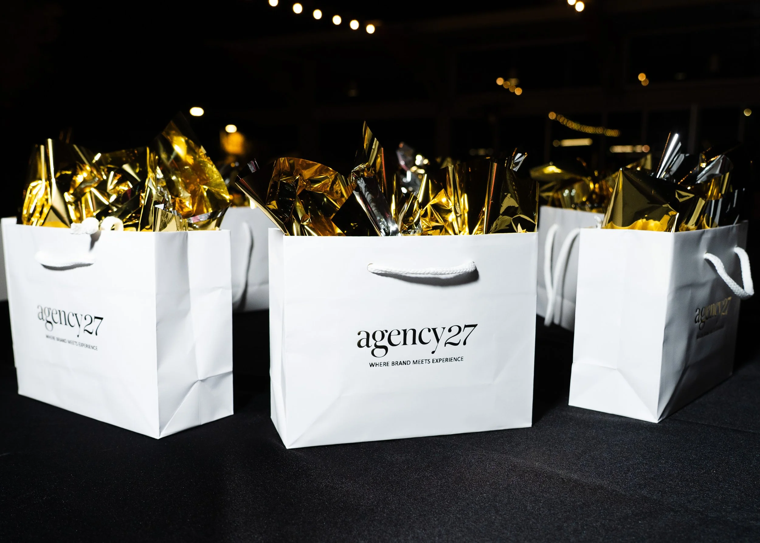 White gift bags with 'agency27' logo and tagline 'Where brand meets experience', filled with gold and silver foil wrapping, placed on a black surface against a dark background.