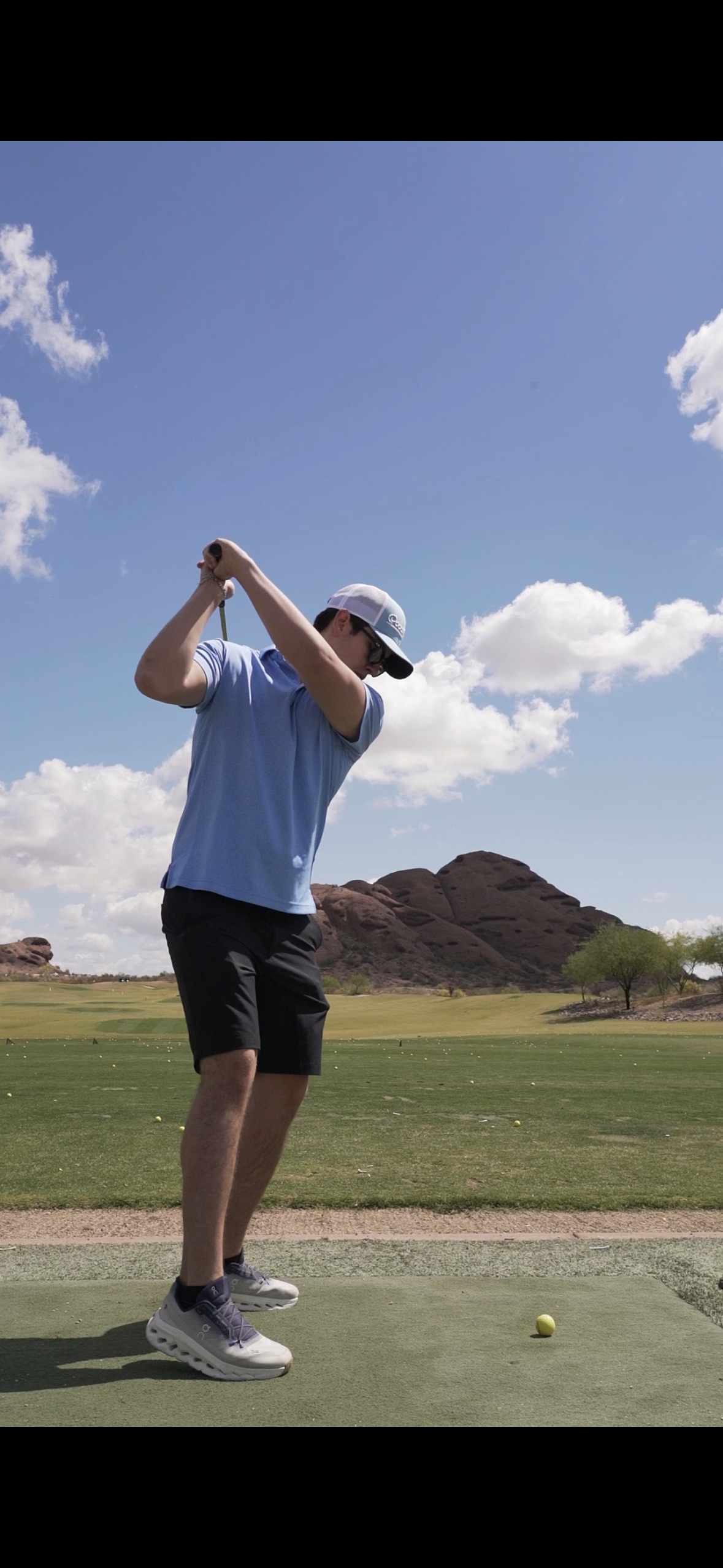 A man on a golf course swinging a golf club; he is wearing a light blue polo shirt, black shorts, white sneakers, sunglasses, and a baseball cap, with a golf ball in front of him and rocky hills in the background under a blue sky with white clouds.