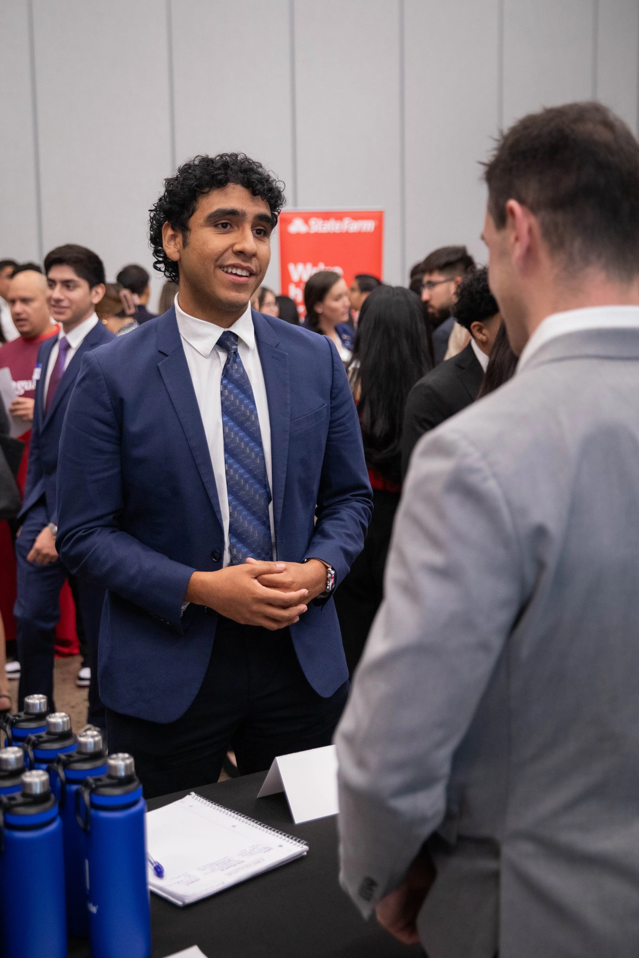 A man in a blue suit and patterned tie talking to another man in a light gray suit at a conference or networking event, with a table of blue water bottles and a notepad in front of them, and a crowd of people in business attire in the background.