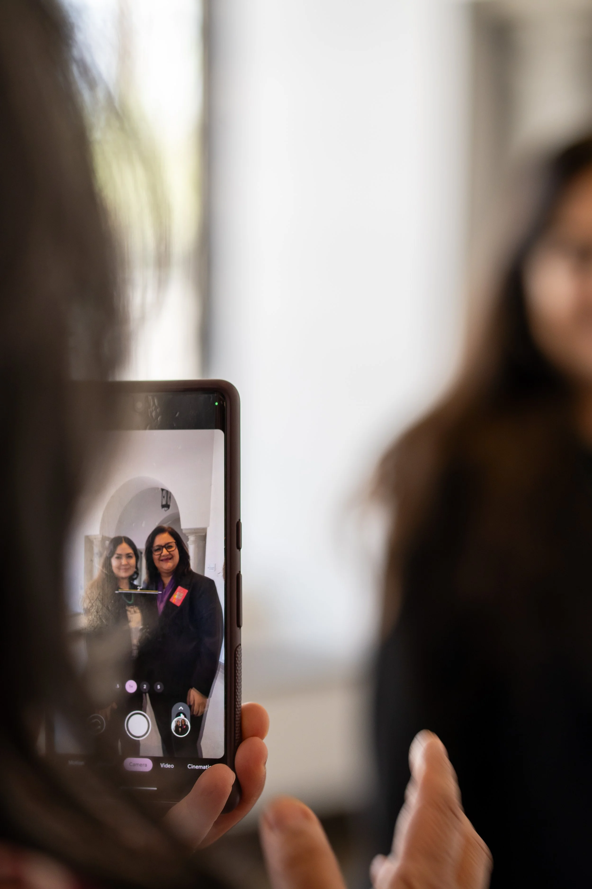 Two women taking a photo in front of a mirror, one woman is smiling and wearing a blazer, the image is seen on a smartphone screen being held by another person.