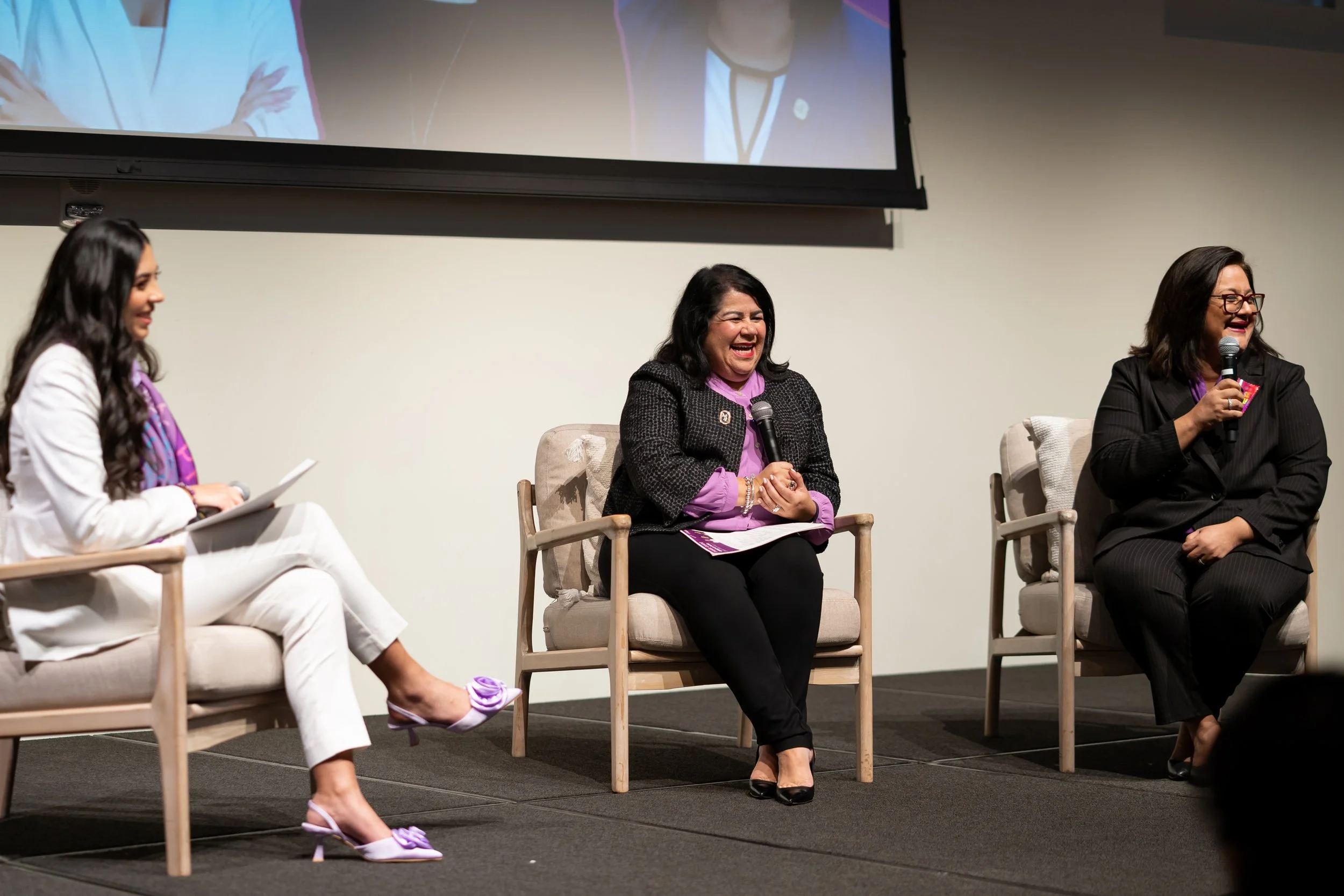 Three women sitting on stage in armchairs, engaged in a panel discussion, with one holding a microphone and laughing, a large screen behind them showing part of the scene.