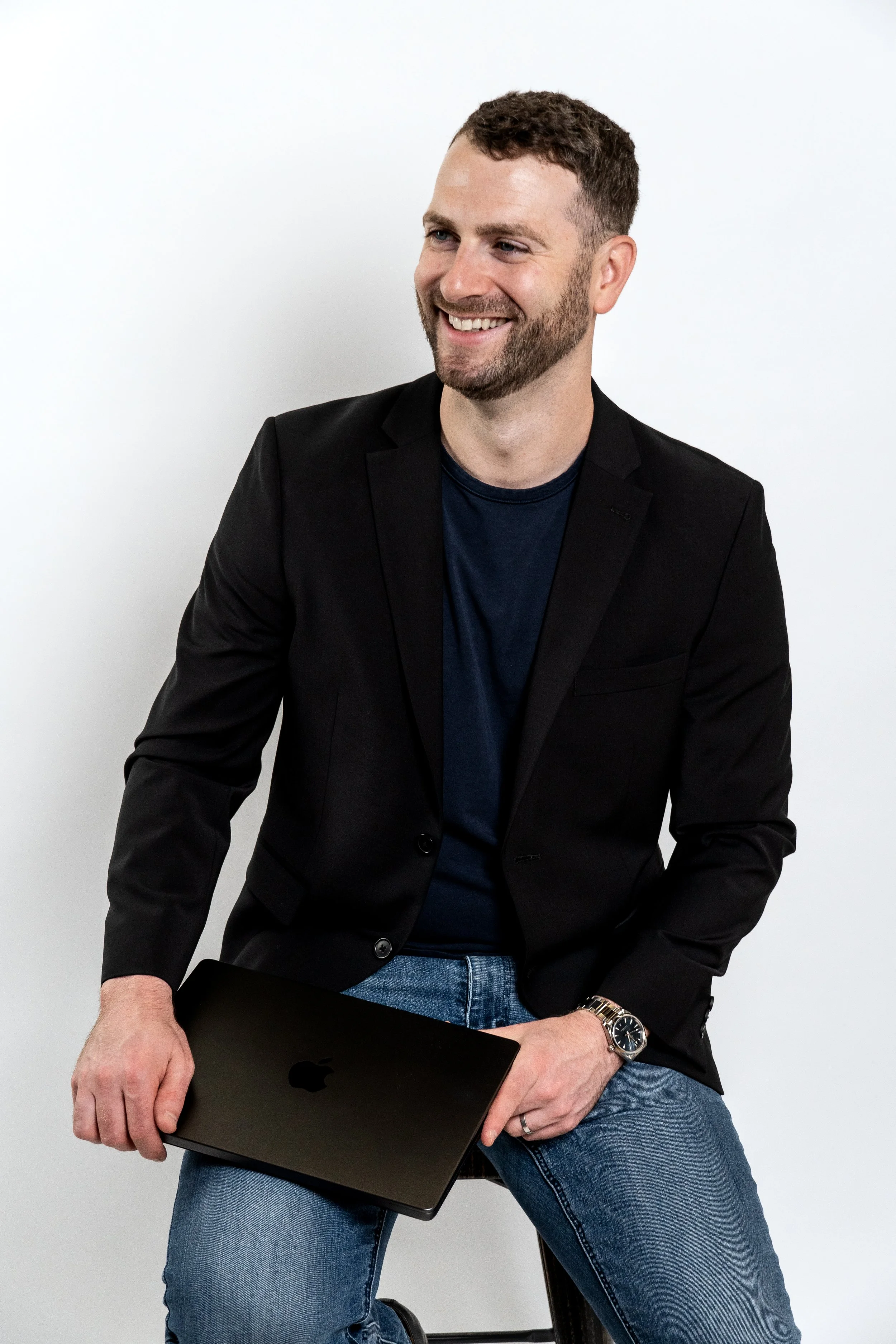 A smiling man with a beard, wearing a black blazer, navy t-shirt, and jeans, sitting on a stool and holding a closed black Apple MacBook in his right hand against a plain white background.