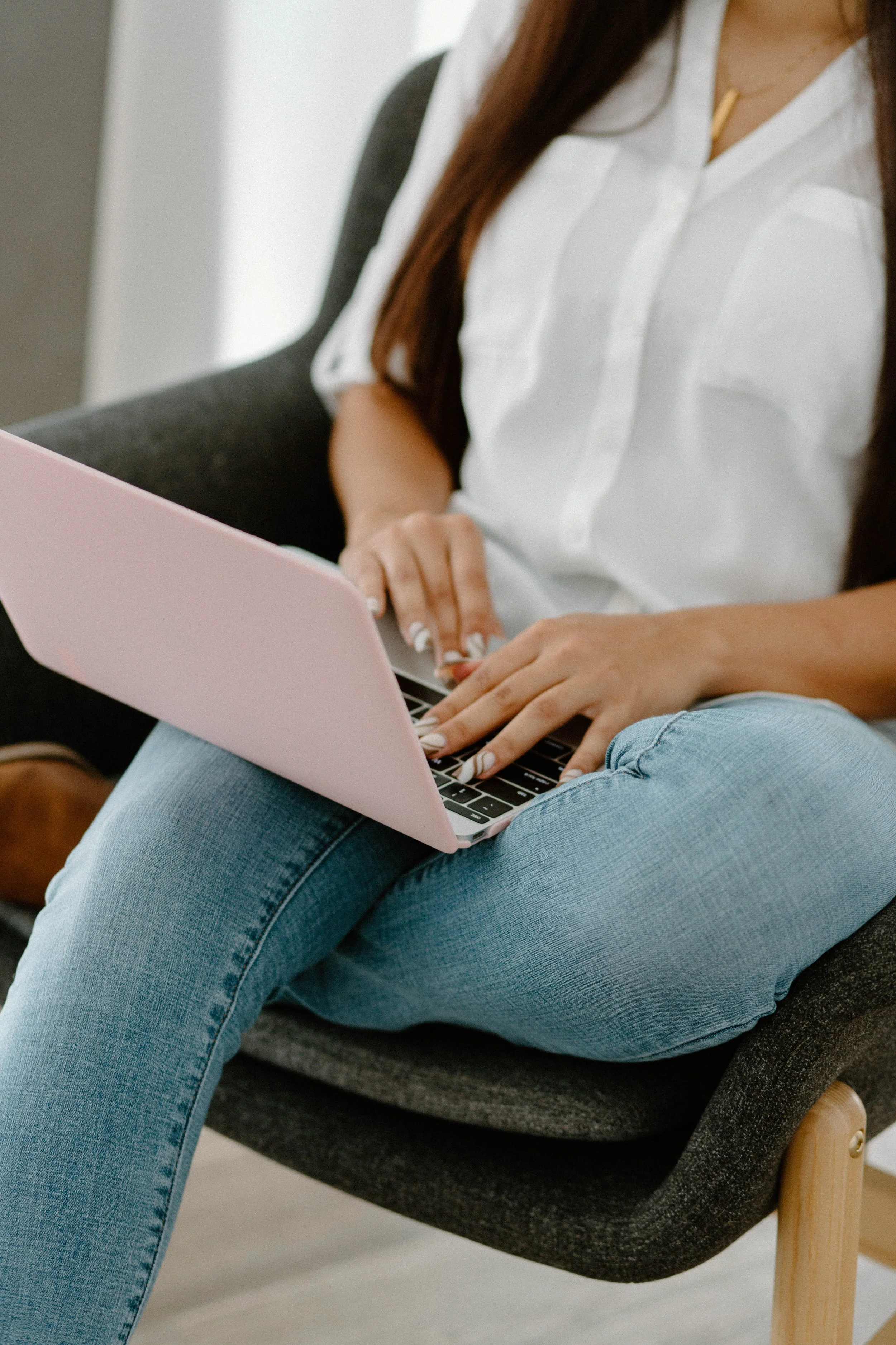 Person sitting on a black chair using a pink laptop, wearing a white shirt and light blue jeans.