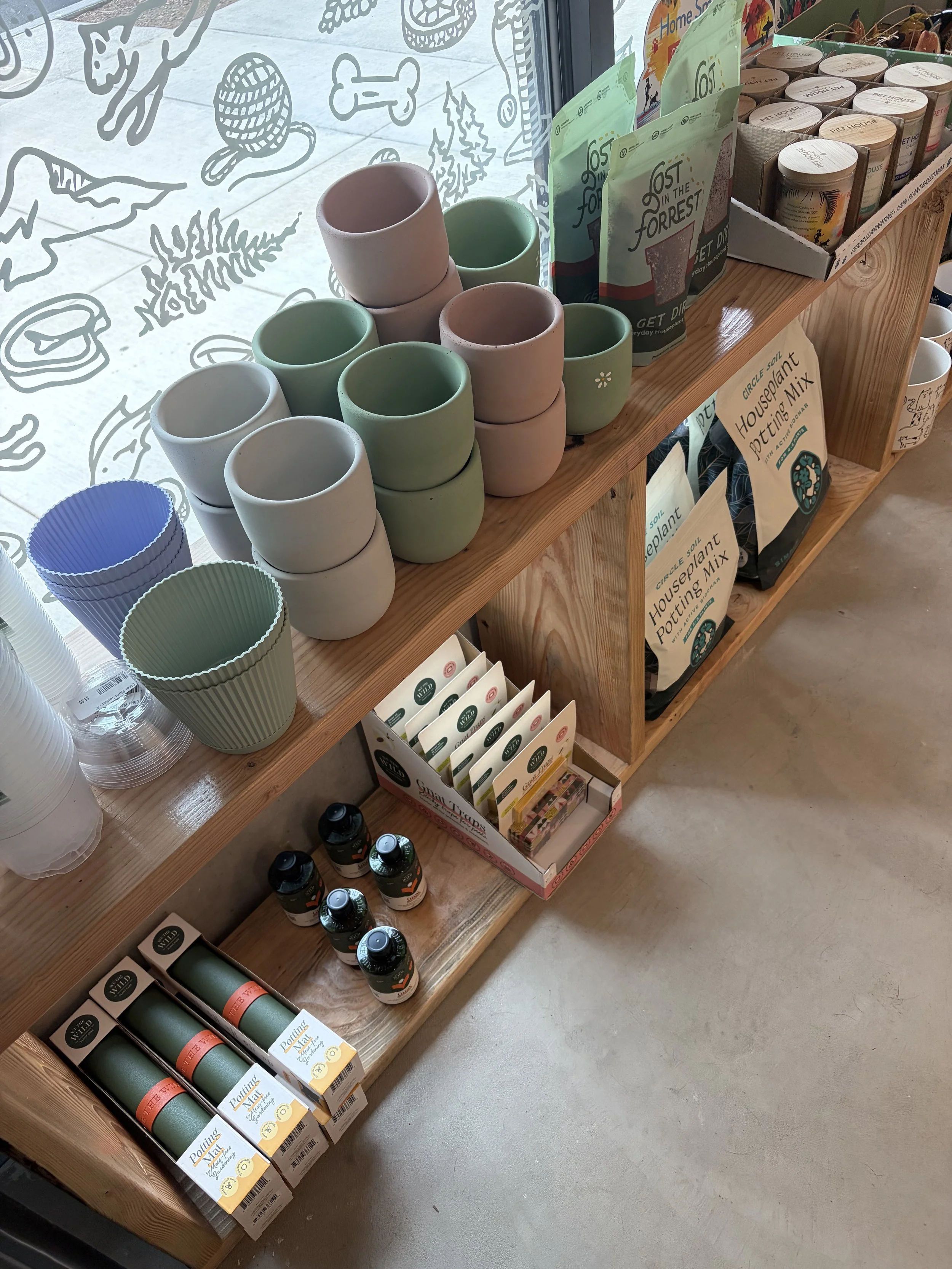 Display of ceramic pots in pastel pink, green, and white on a wooden shelf, alongside pet-related products such as pet food and supplements in a store.