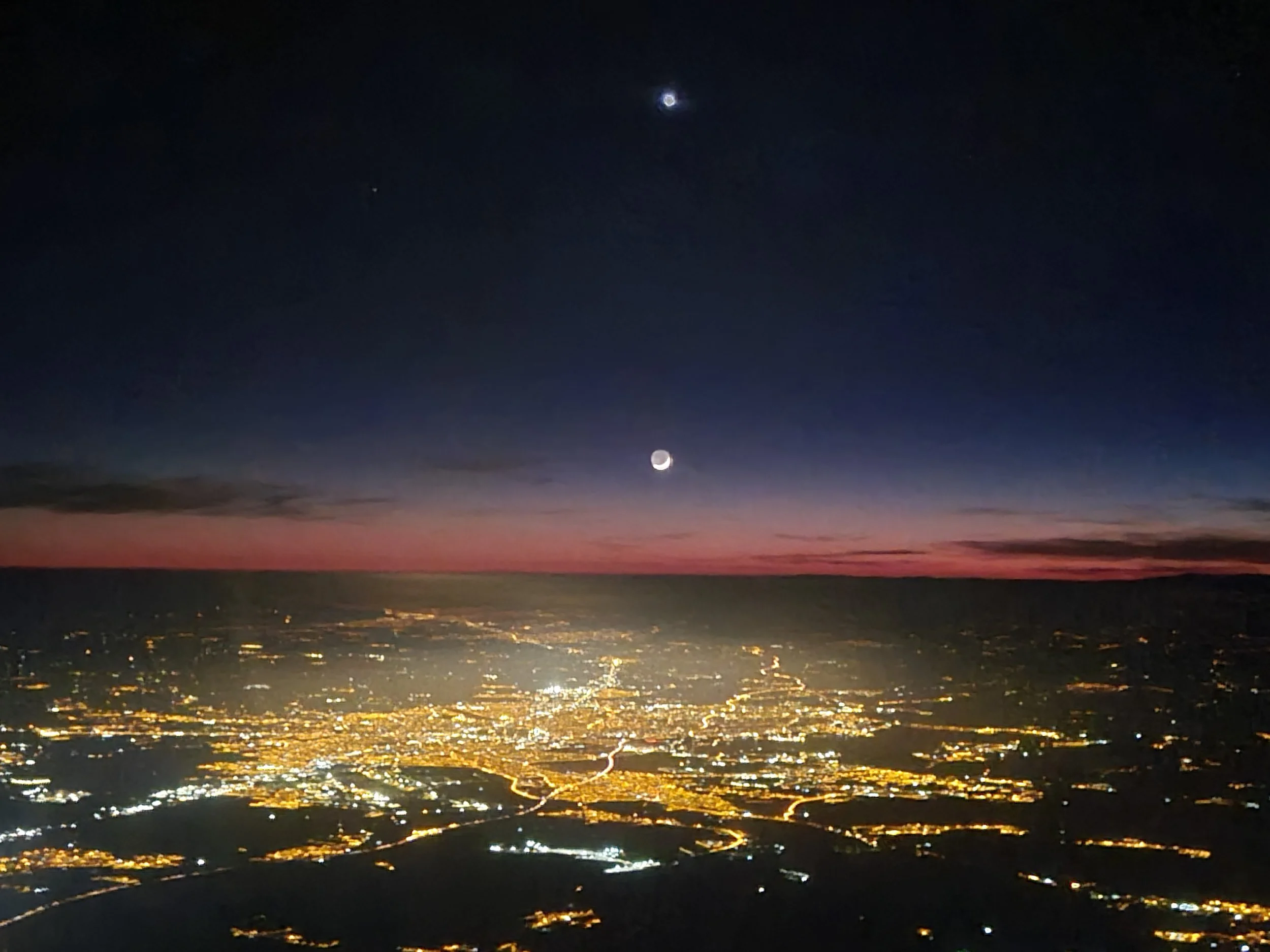 Aerial night view of Ankara, Türkiye with illuminated streets and buildings, a sunset horizon, and a dark sky with the moon and a bright planet or star visible.