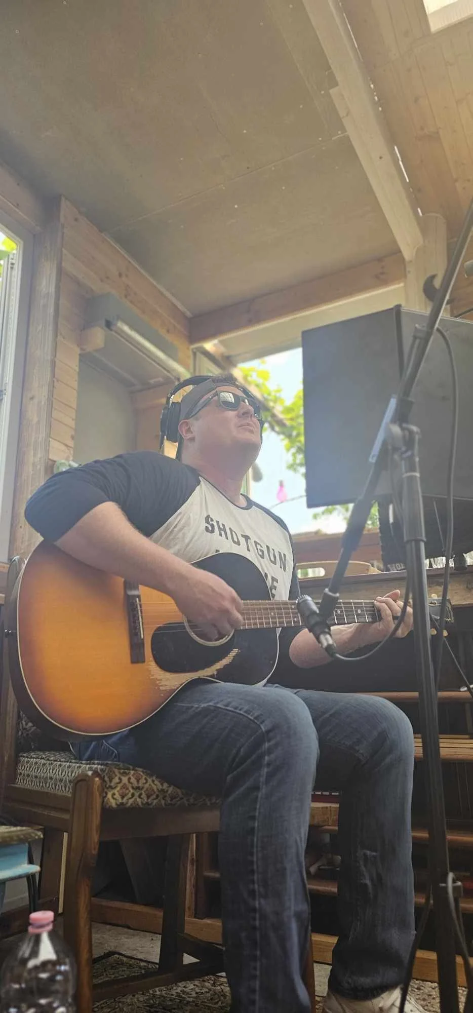 Man playing acoustic guitar, wearing sunglasses and a t-shirt with 'SHOTGUN WILLIE' printed on it, seated indoors with wooden walls and ceiling, music equipment in front, and sunlight coming through window. Live recording of Tumbleweed Blues.