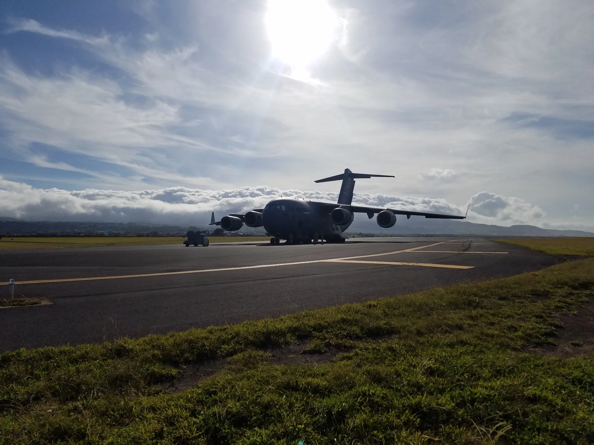 A large military cargo airplane (C-17A Globemaster) on a US Air Force airport tarmac in the Azores with a cloudy sky and the sun overhead.
