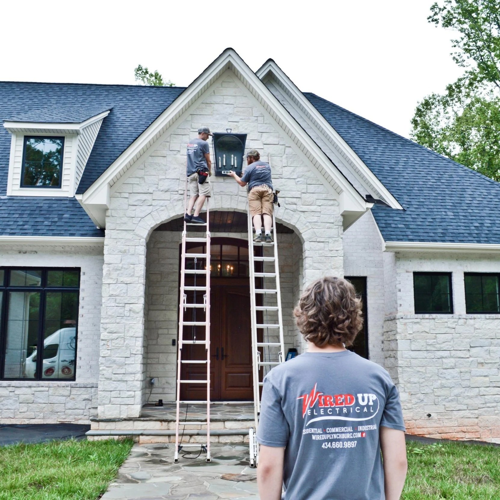 Professional electricians on ladders installing outdoor electrical outlets on a home exterior, following safety standards and proper wiring practices.