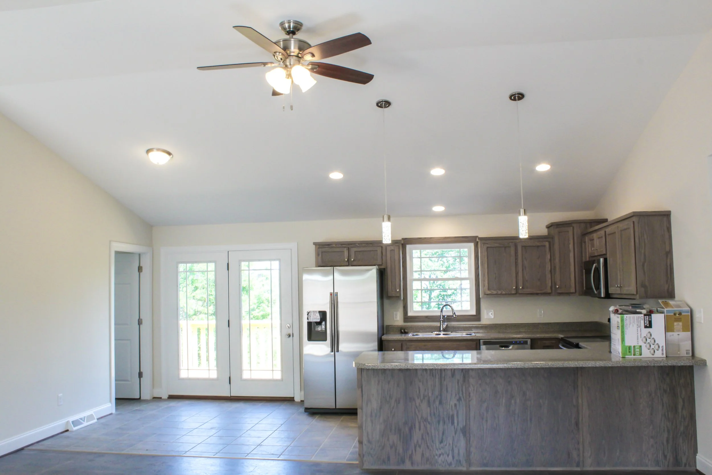 Kitchen with gray wooden cabinets, stainless steel refrigerator, double sink, windows, tile flooring, two pendant lights, ceiling fan with three lights, and double glass doors leading outside.
