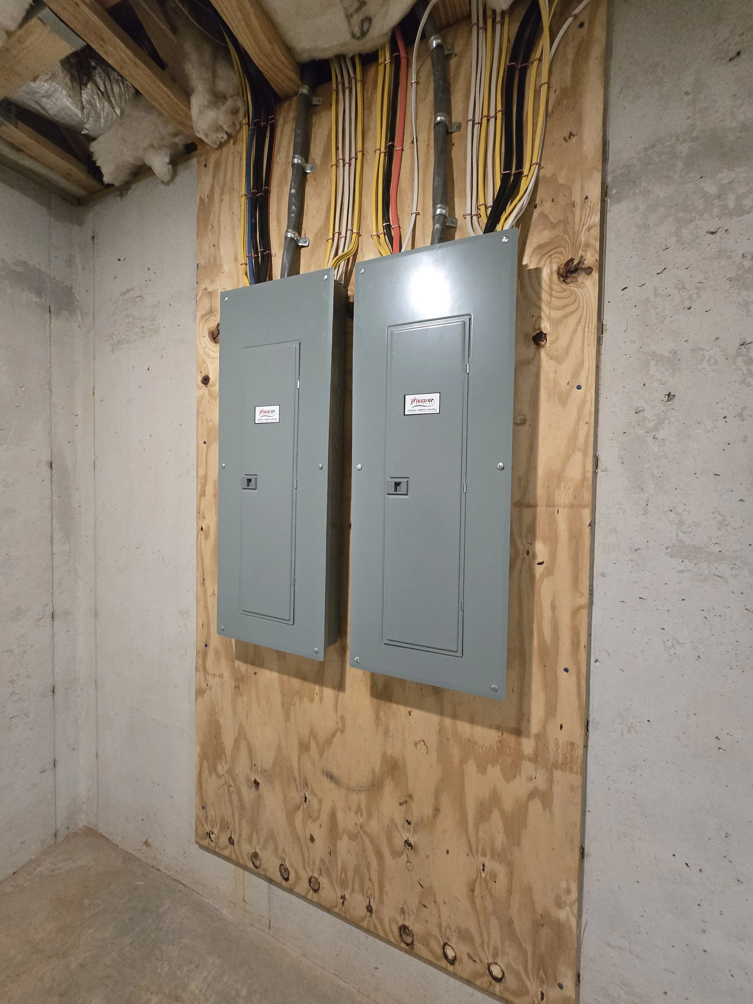 Two gray electrical breaker boxes mounted on a wooden board in a basement with exposed insulation and concrete walls.