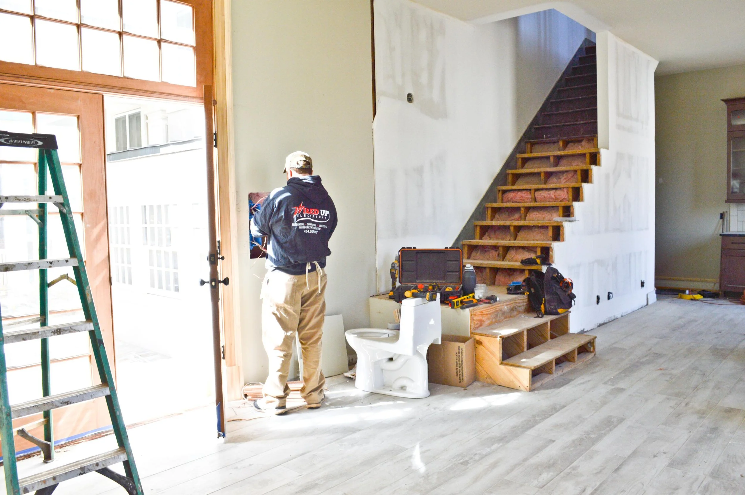 A worker inside a house under renovation, facing away, working near a door opening to the outside, with tools on a ledge next to a staircase that is under construction, and a white toilet in the room.