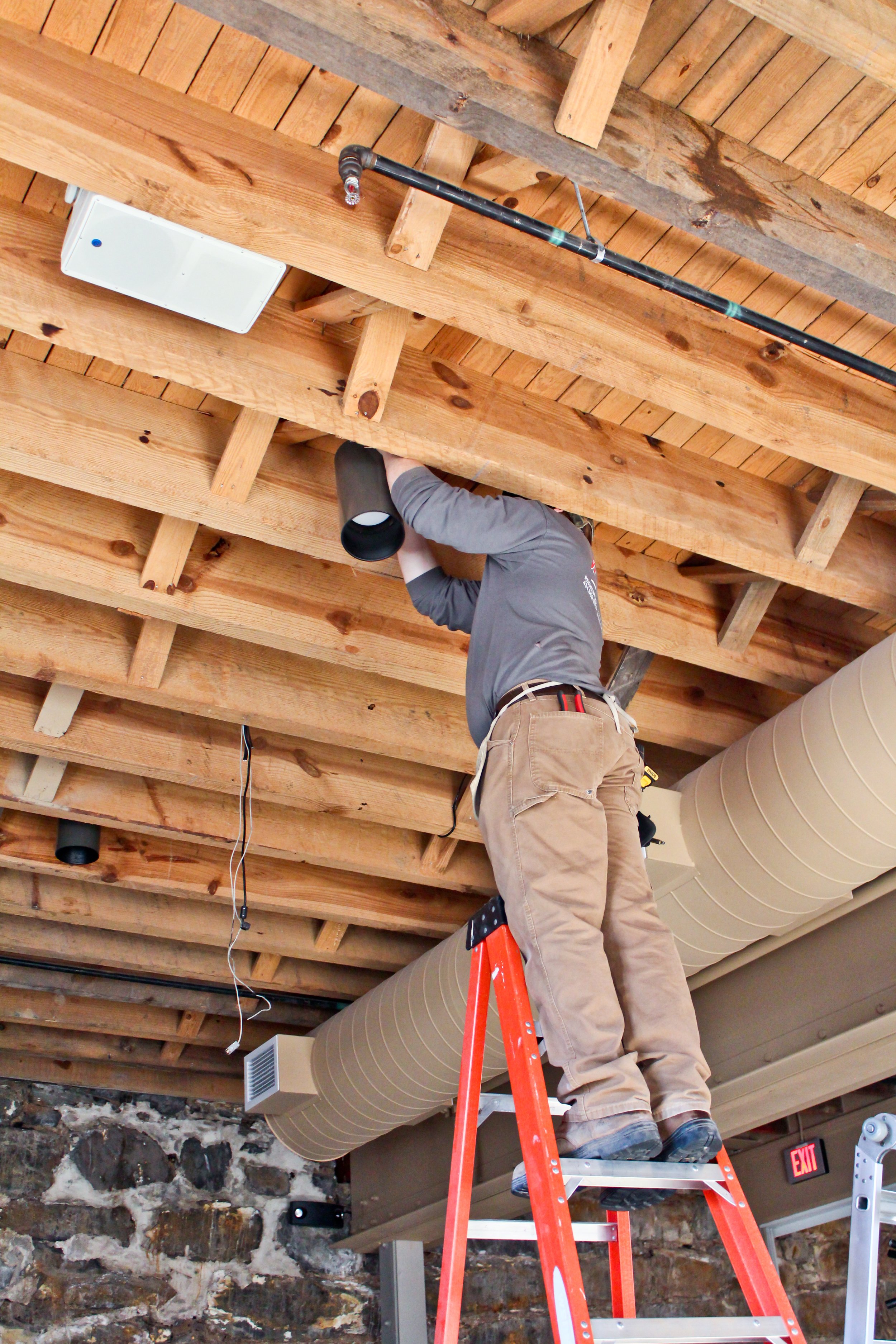 A person on an orange ladder installing or inspecting the building's wooden ceiling framework, with HVAC ductwork and electrical wiring visible.