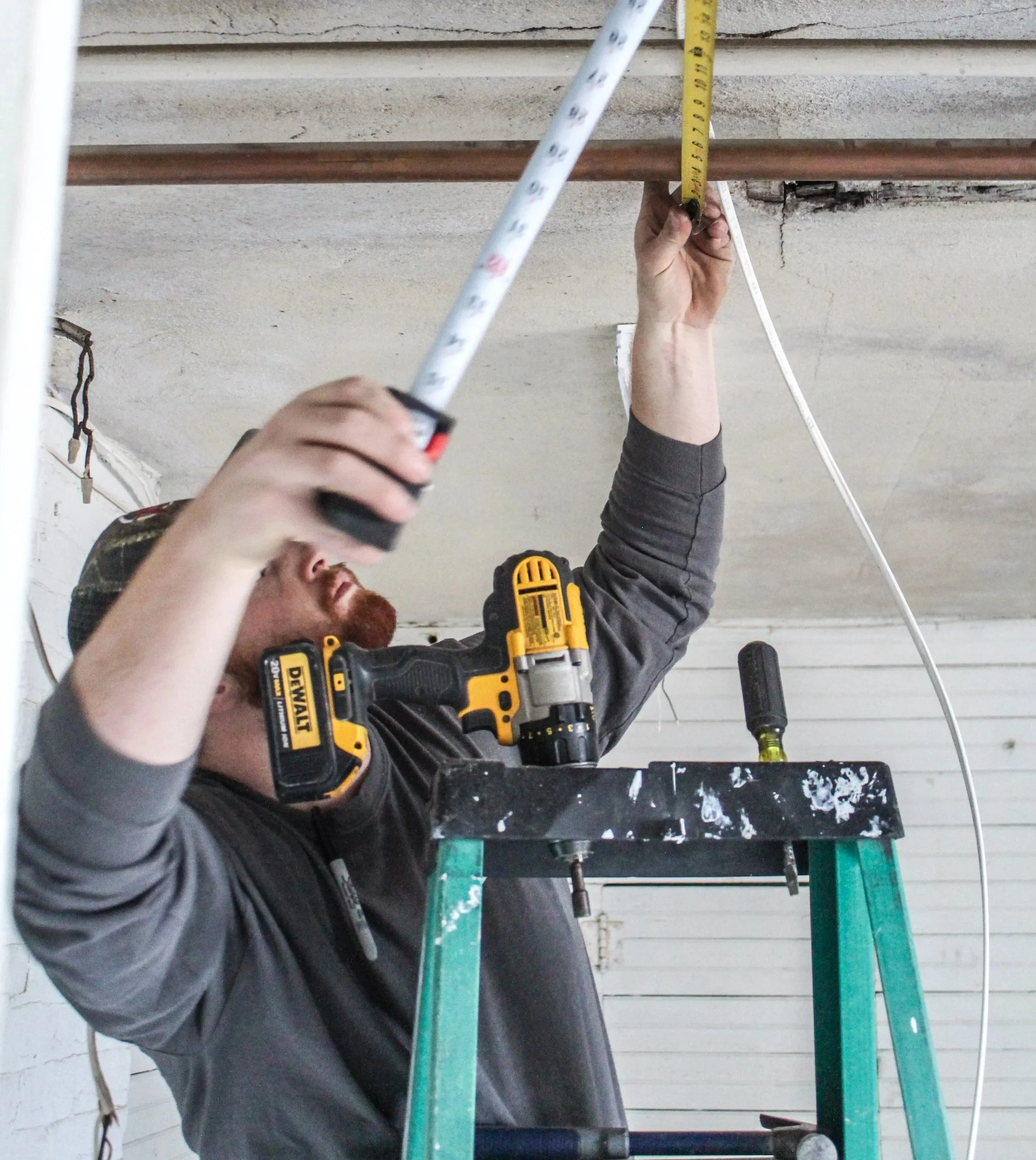 Man on a green ladder measuring the ceiling with a tape measure, using a cordless drill strapped to his shirt, in a construction or renovation setting.