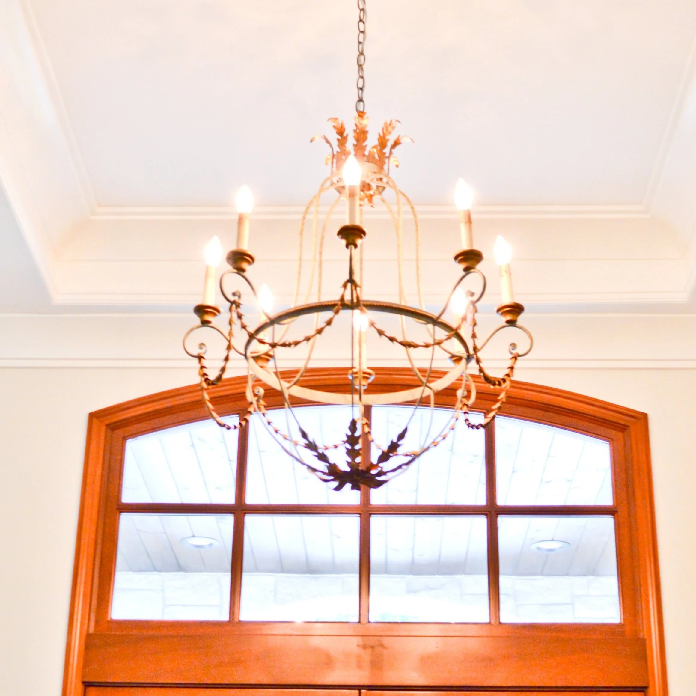 Decorative chandelier hanging from ceiling, illuminated above a large arched window inside a bright residential interior.