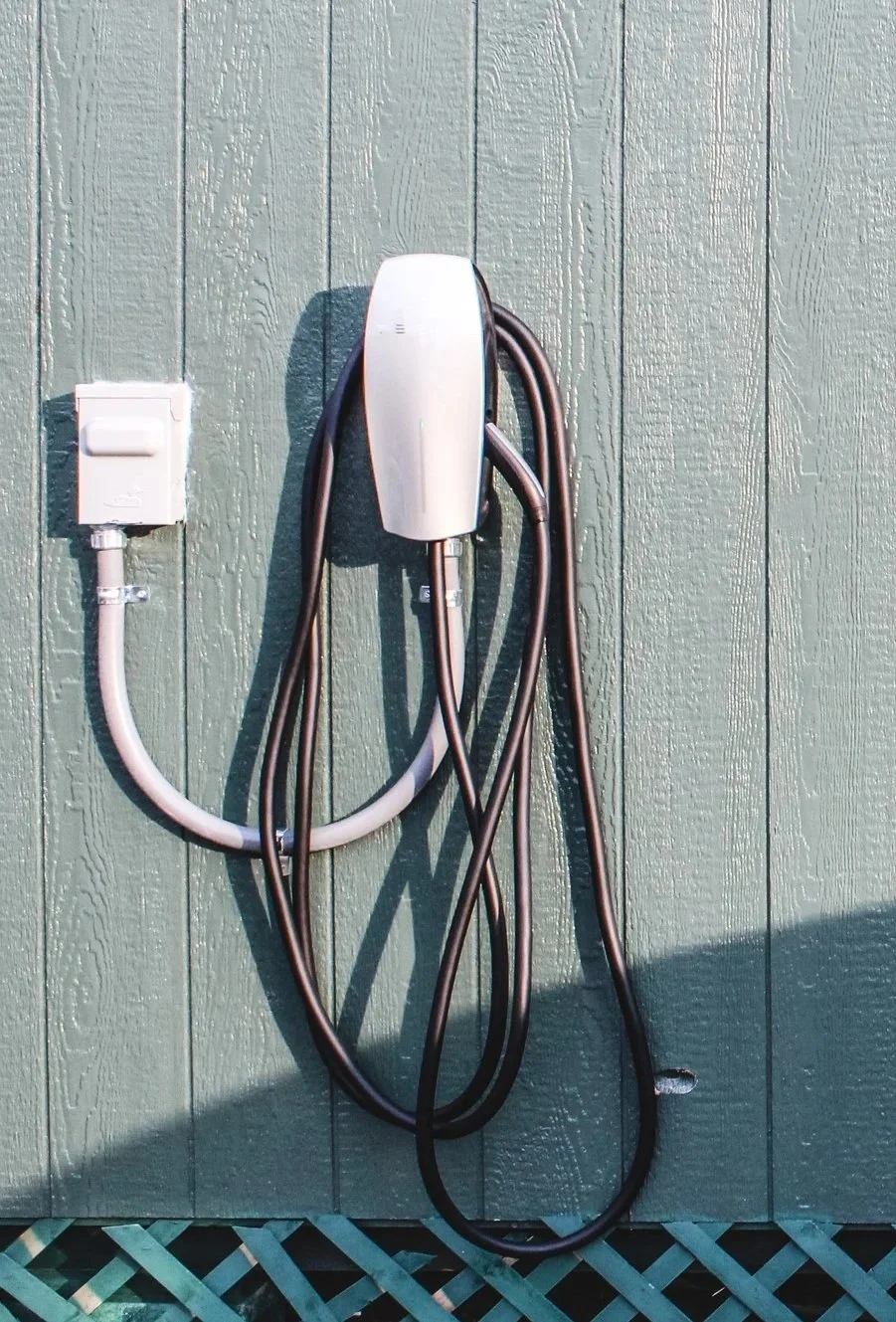 An electric vehicle charging station mounted on a light green wooden exterior wall, with black and white charging cords hanging from it.