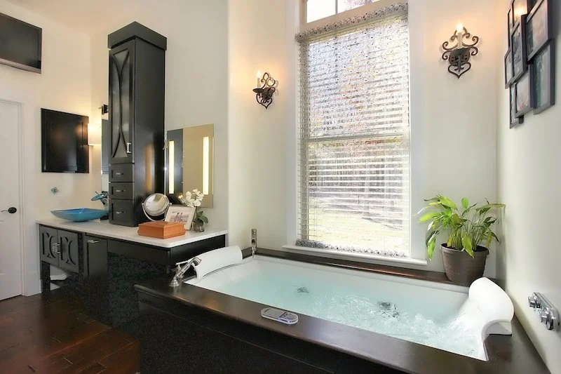 A bathroom with a large soaking tub filled with water, a black vanity with a blue sink and a mirror, a tall black cabinet, a window with blinds, a potted plant, and wall-mounted lights.