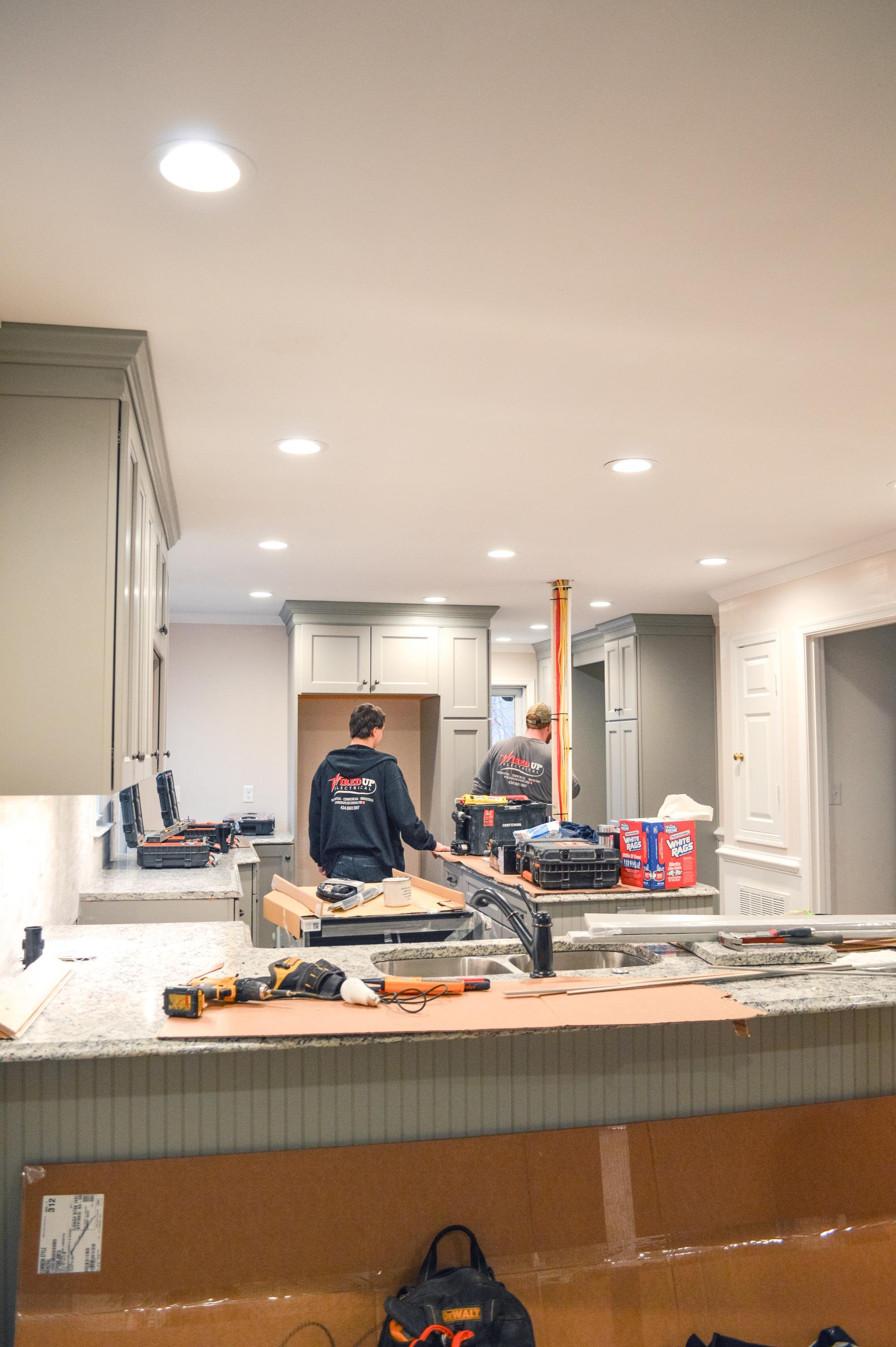 Two workers in black hoodies working on kitchen renovation with gray cabinets and granite countertops, various tools and supplies on the counter.