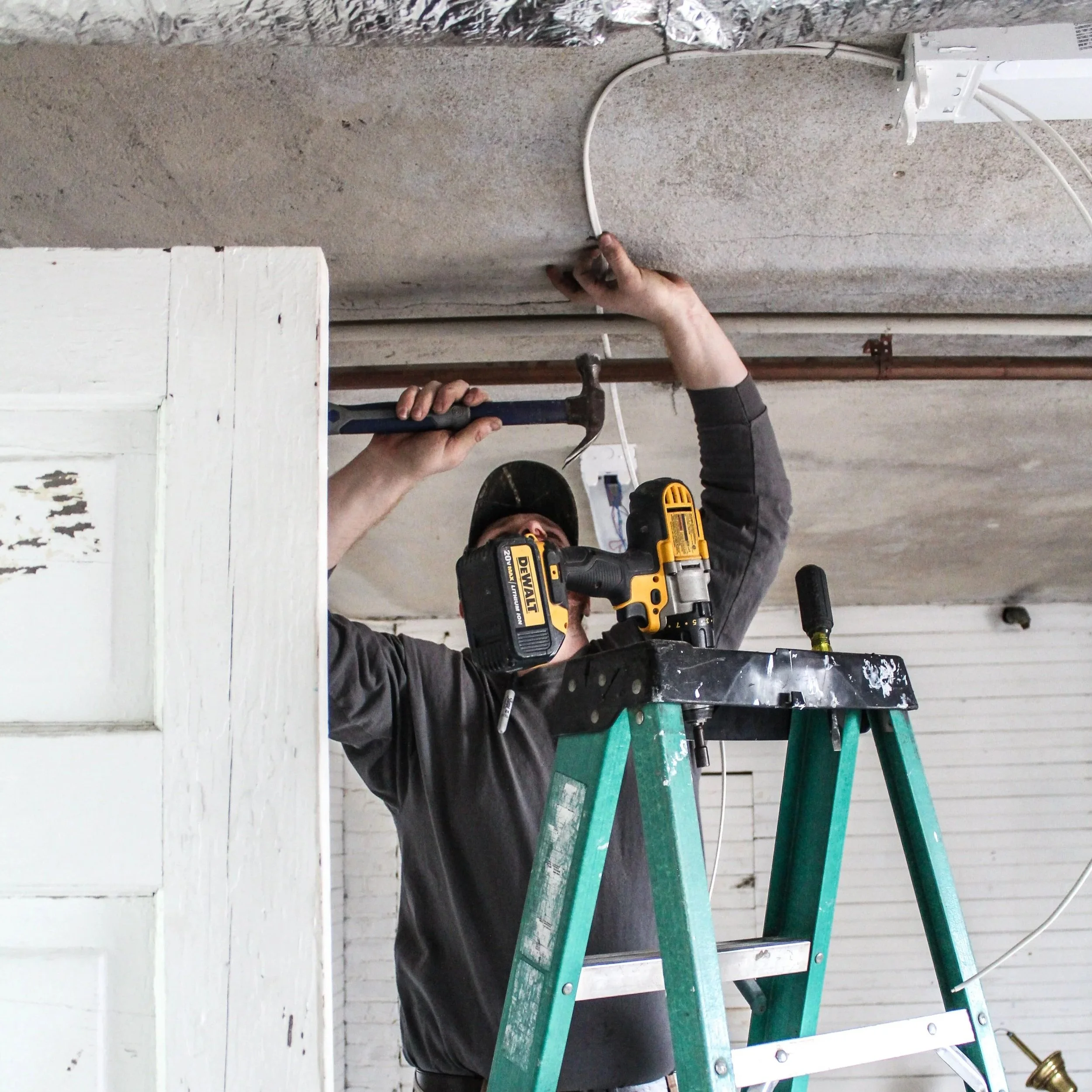 Electrician standing on ladder, using hammer and drill to secure ceiling electrical wiring inside a residential space.