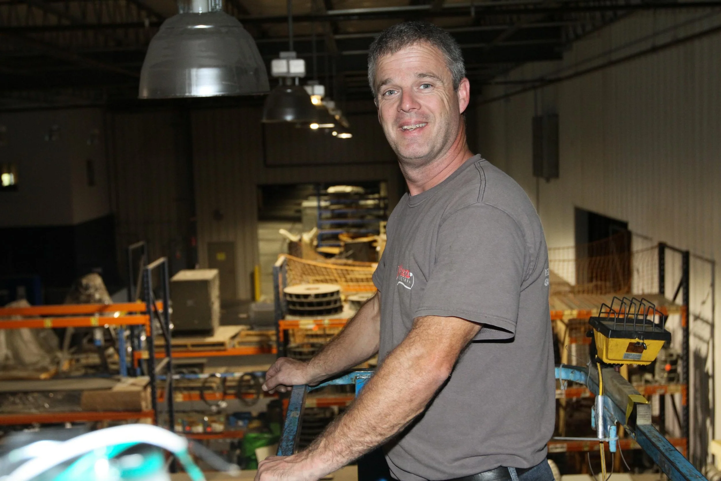A man standing on a ladder in an industrial warehouse, smiling and looking at the camera, with shelves and equipment in the background.