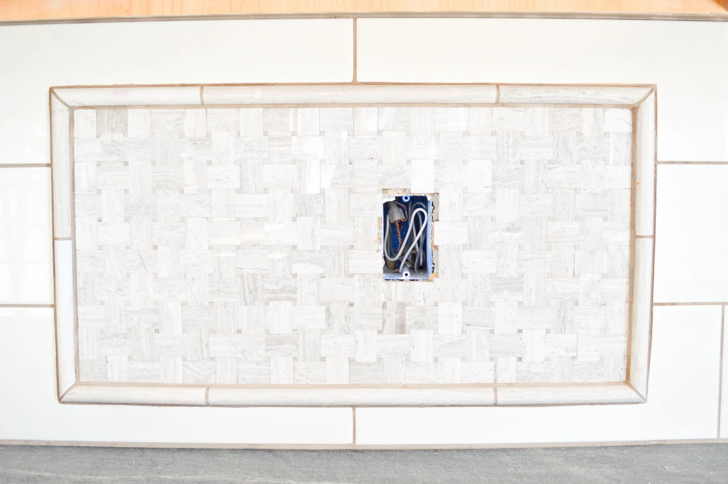 Wall with a central electrical outlet box, wires inside, and a raised tile border surrounding the outlet area.