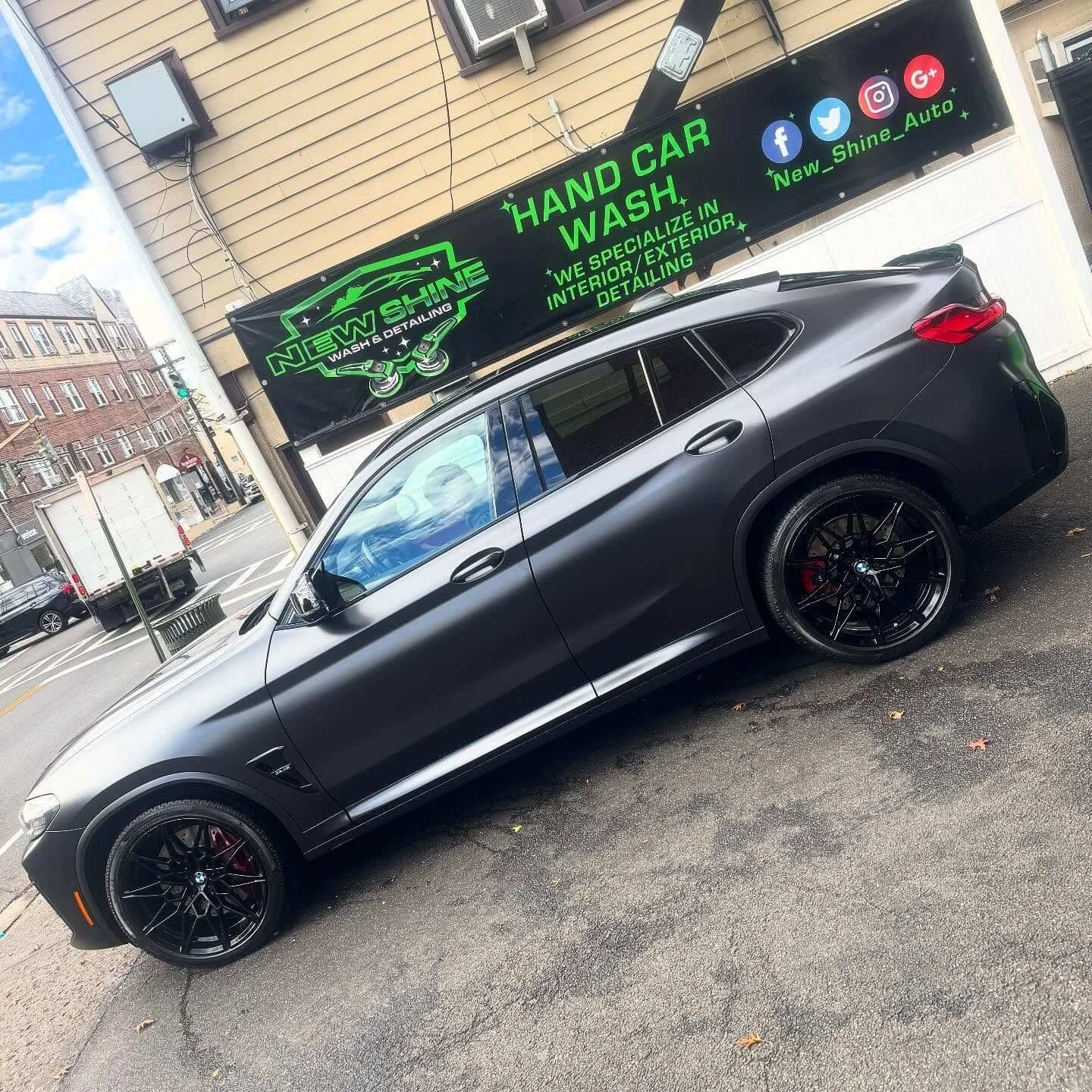 A black BMW SUV parked outside a car wash and detailing business with a green and black sign showcasing their services and social media icons.