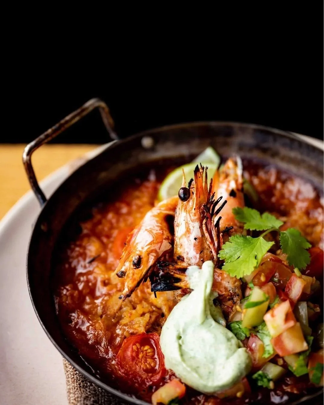 A close-up of a shrimp dish in a black skillet, garnished with cilantro, lime, cherry tomatoes, and a dollop of green sauce.