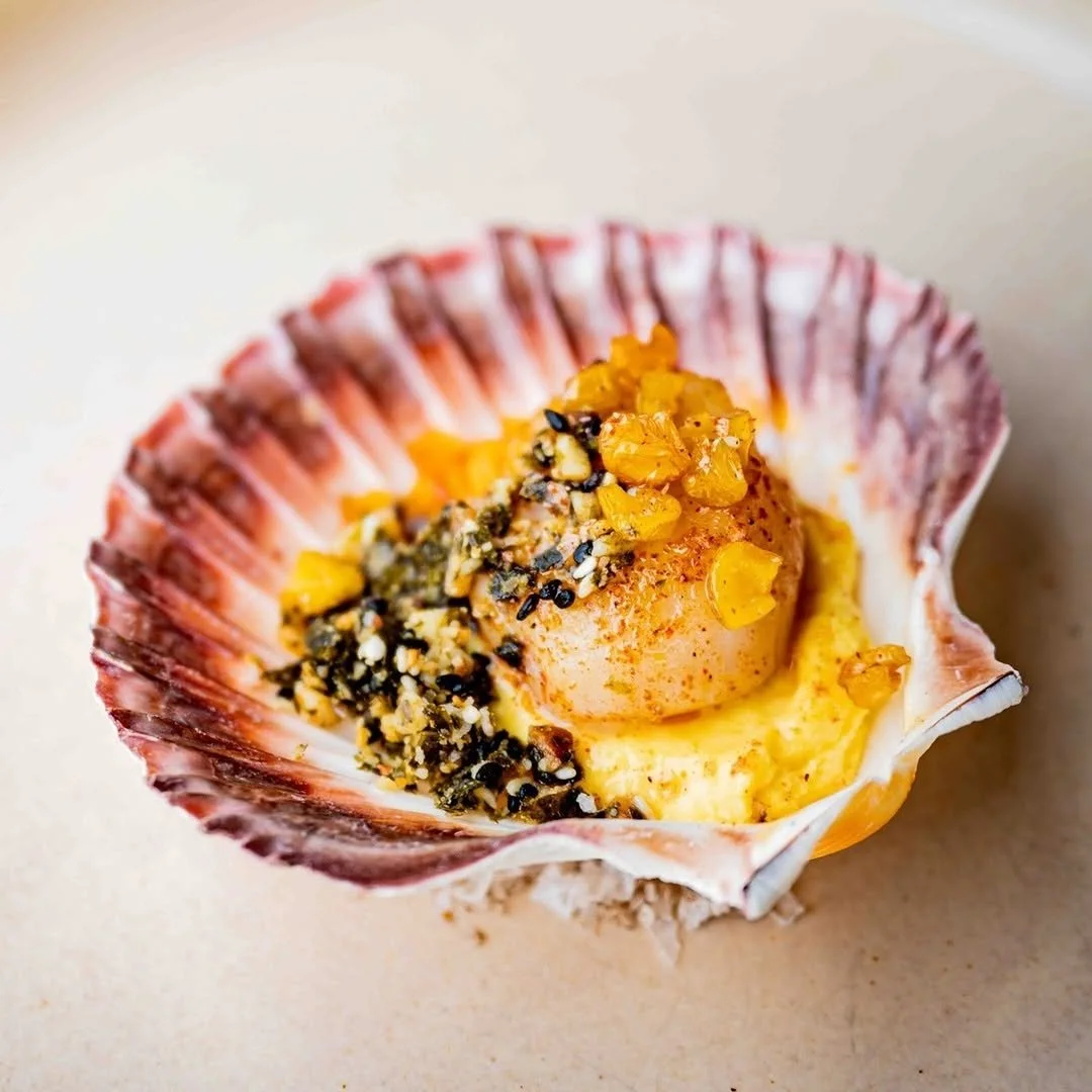 Sea shell with scoop of ice cream topped with crushed nuts and yellow fruit pieces, on a beige background.