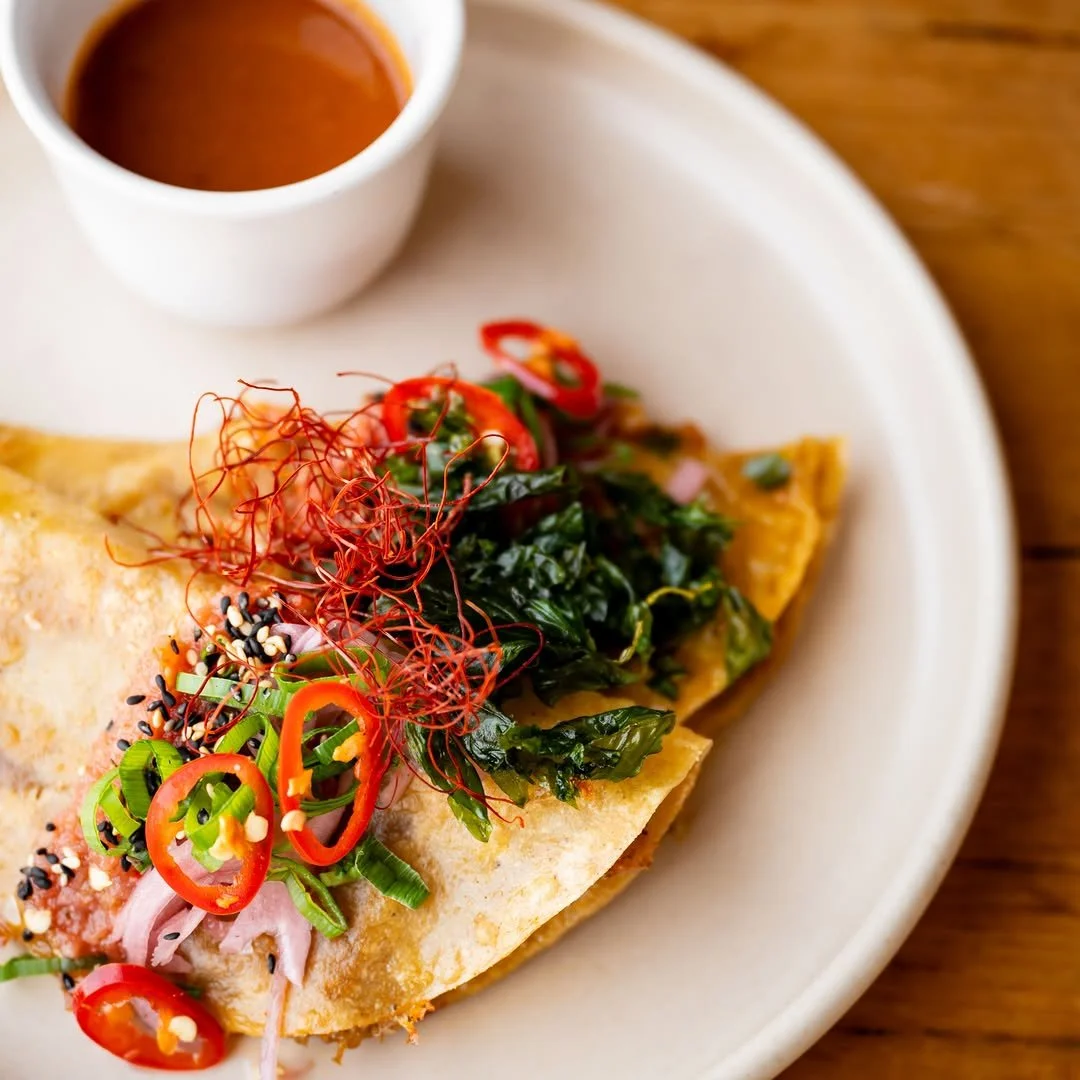 Taco on a white plate topped with sliced red chili peppers, chopped green onions, black and white sesame seeds, cooked leafy greens, and thin red chili threads, with a small cup of sauce in the background.