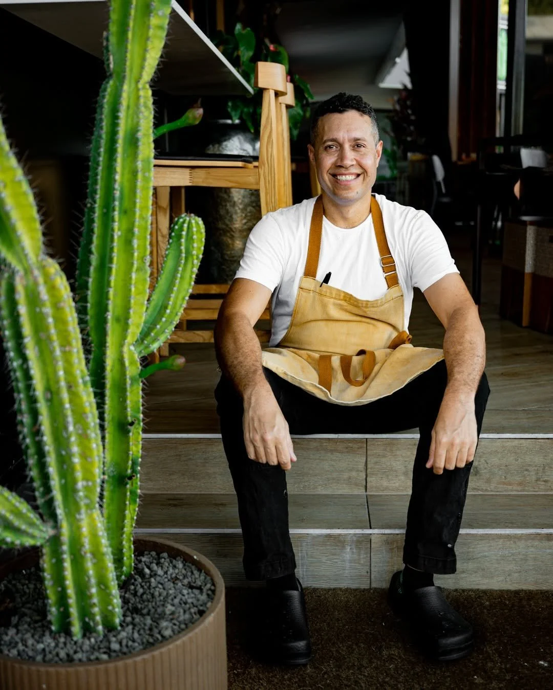 A smiling man sitting on steps in a plant shop, wearing a white t-shirt, black pants, black shoes, and a beige apron. There is a tall cactus in the foreground and indoor plants and furniture in the background.