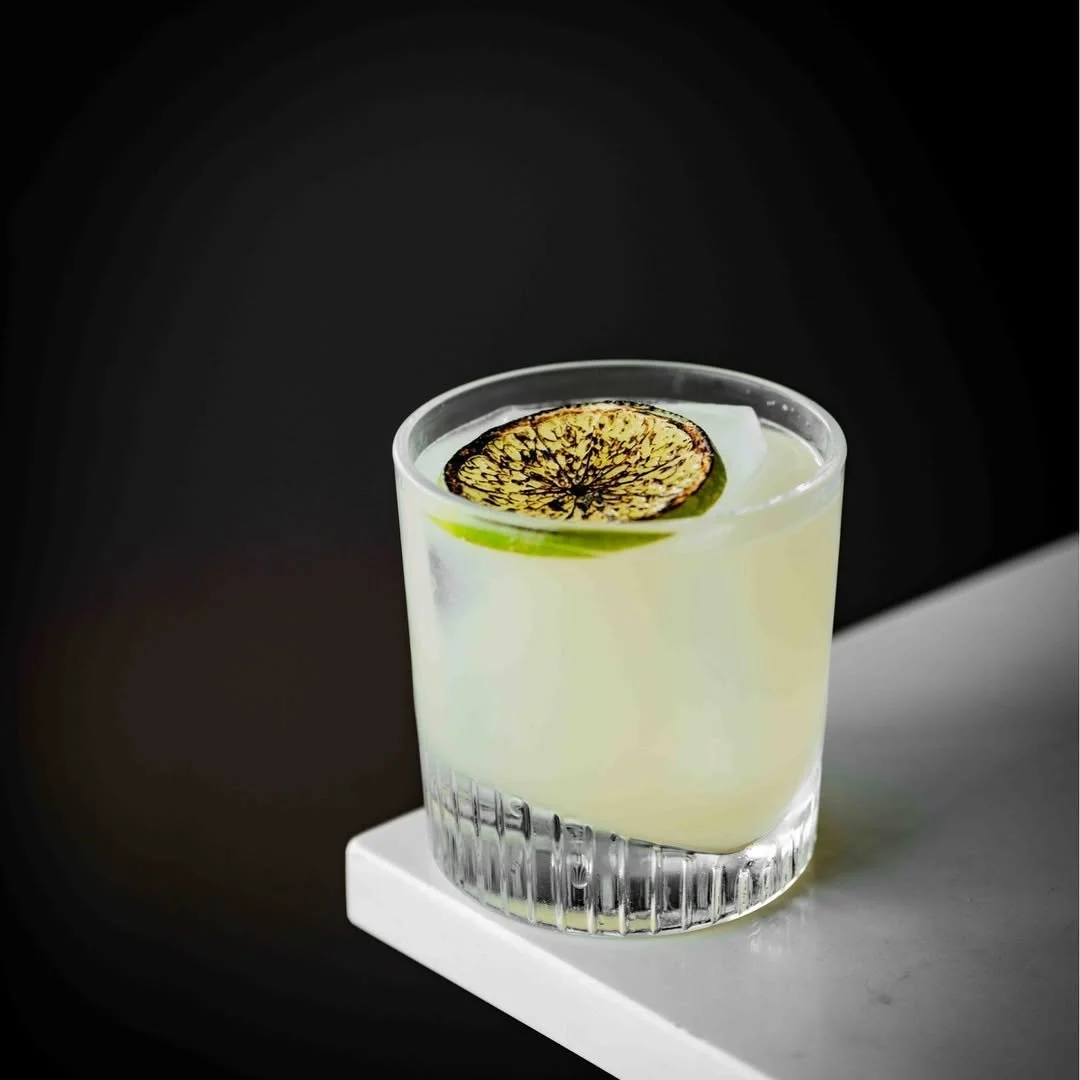 A clear glass of a light-colored cocktail garnished with a thin, charred lime wheel, placed on a white surface against a dark background.