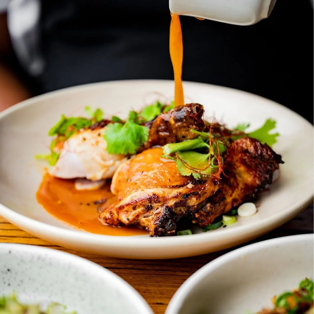 Pouring sauce over grilled chicken thigh garnished with cilantro and green onions on a white plate.