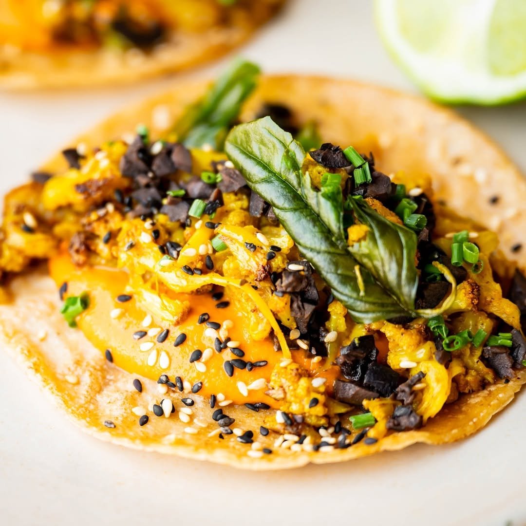 Close-up of a breakfast taco with scrambled eggs, cheese, black beans, green chives, and a basil leaf, served on a corn tortilla with lime wedges in the background.