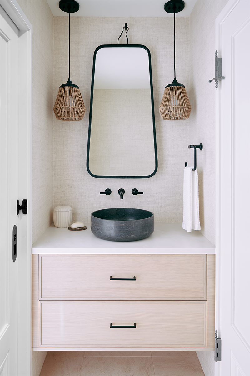 This single vanity with a natural stone vessel sink is located within a shared powder room/toilet room for the primary bathroom. In here we create a peaceful interior through a light linen feel vinyl wallpaper and fun basket pendants.