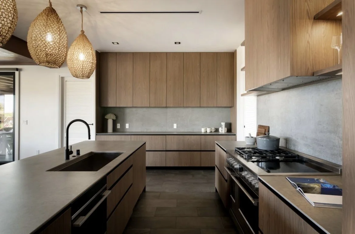 A bar sits along the perimeter of the kitchen featuring mid tone laminate millwork, dark Dekton countertops and backsplashes, & high end Wolf appliances.