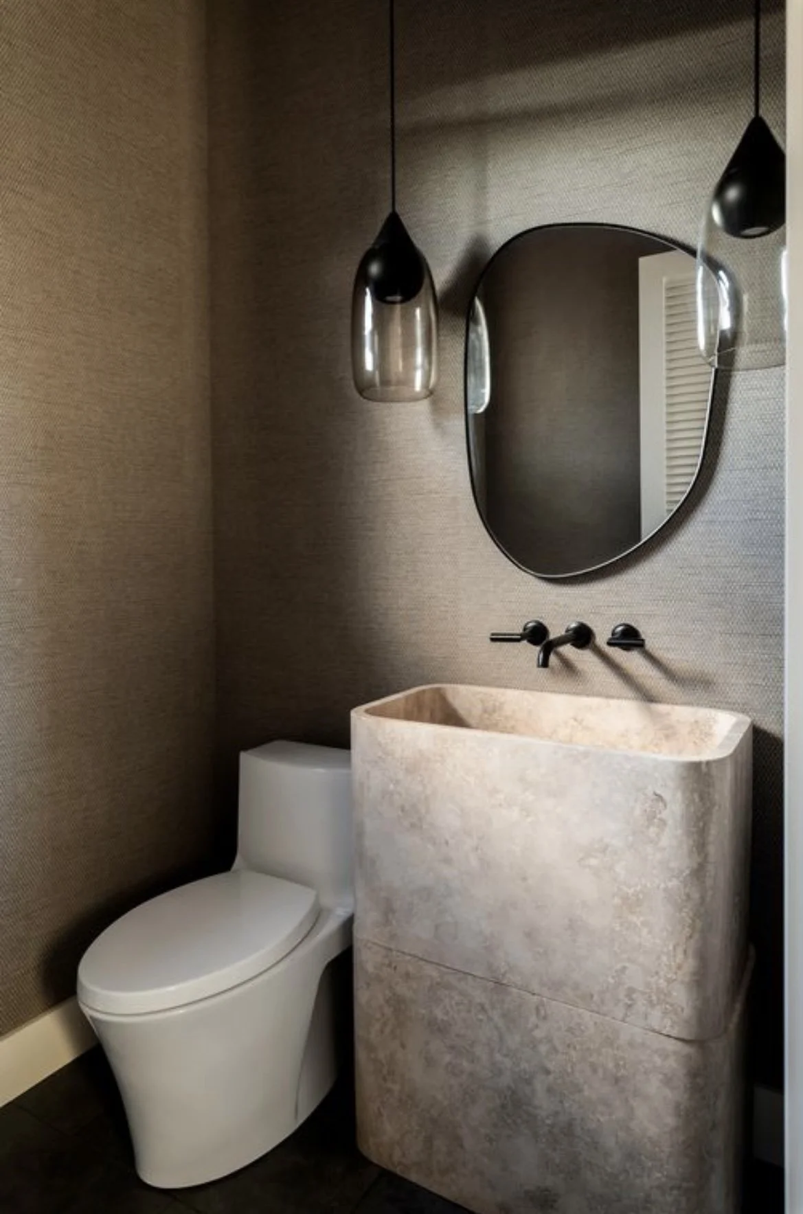 A powder room with a solid stone pedestal sink, black plumbing fixtures and lighting, & a light weaved grasscloth wallpaper.