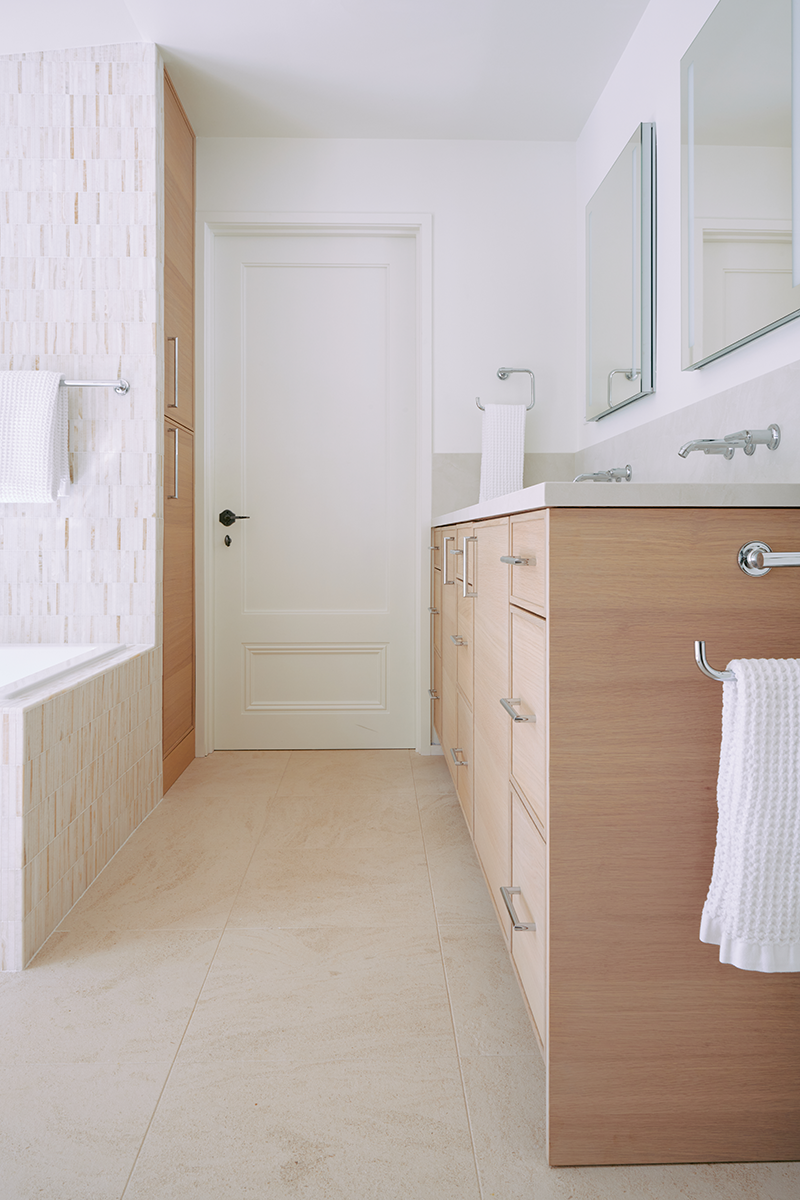 With a tall linen closet tucked away, this initial view into this primary bathroom is stunning and light. The renovation included cabinetry, a new tub and shower, and updated natural stone flooring throughout.