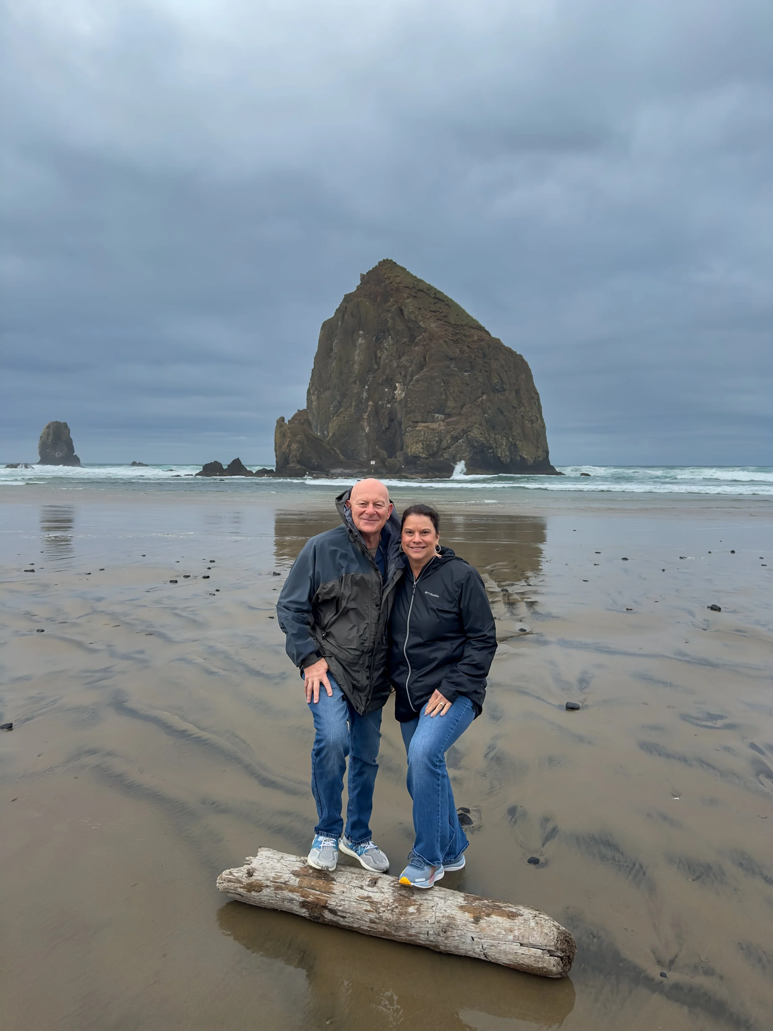 A man and woman standing on a log on a wet beach with large sea stacks in the background, under an overcast sky.