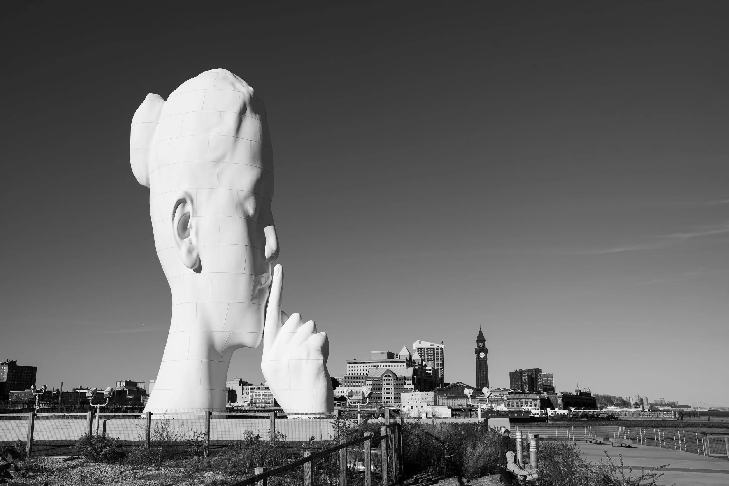 A large white sculpture of a human head with a finger to lips, standing in an urban area with city buildings in the background.